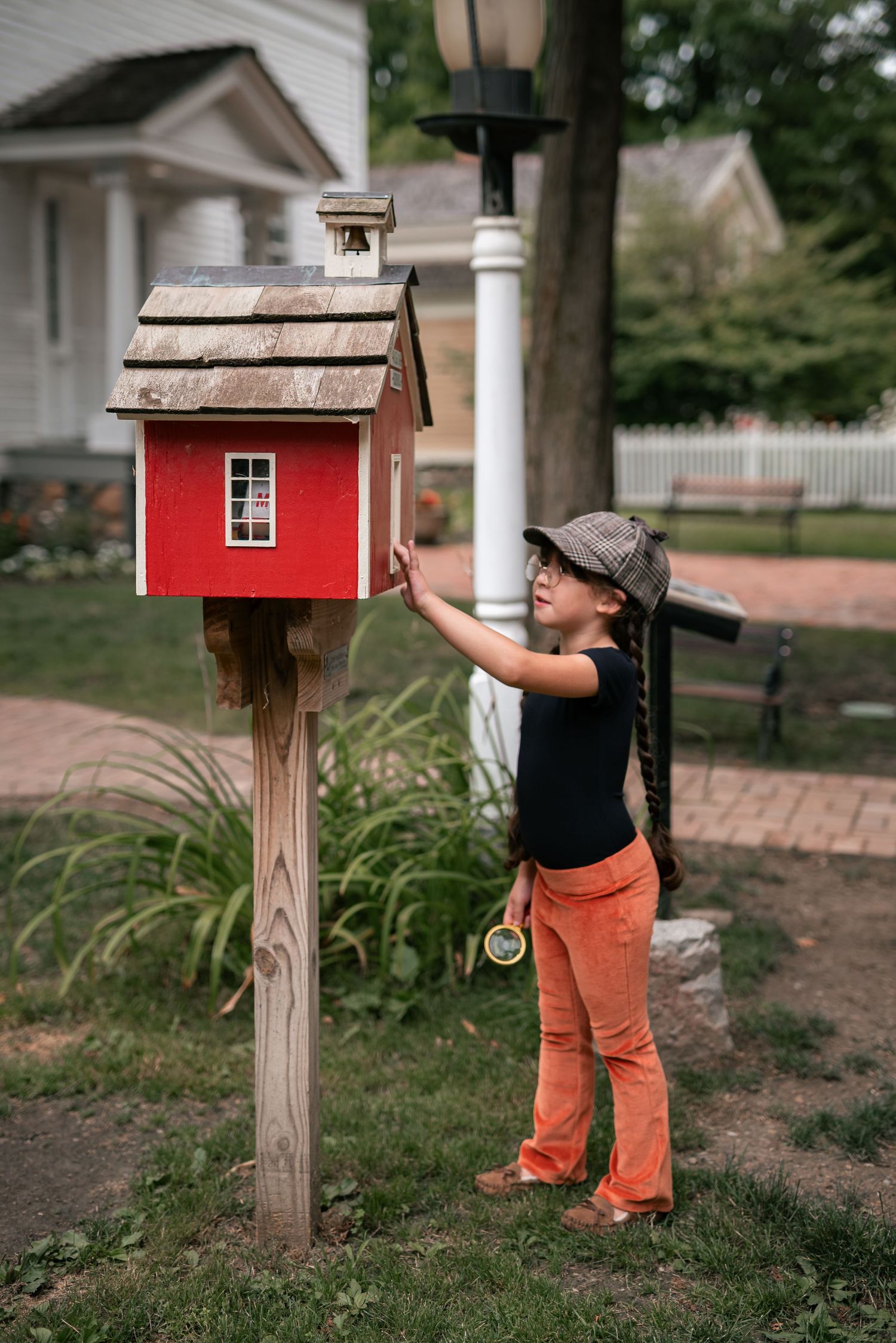 Young girl in orange pants and newsboy cap reaching up to a red miniature house on a wooden post outdoors.
