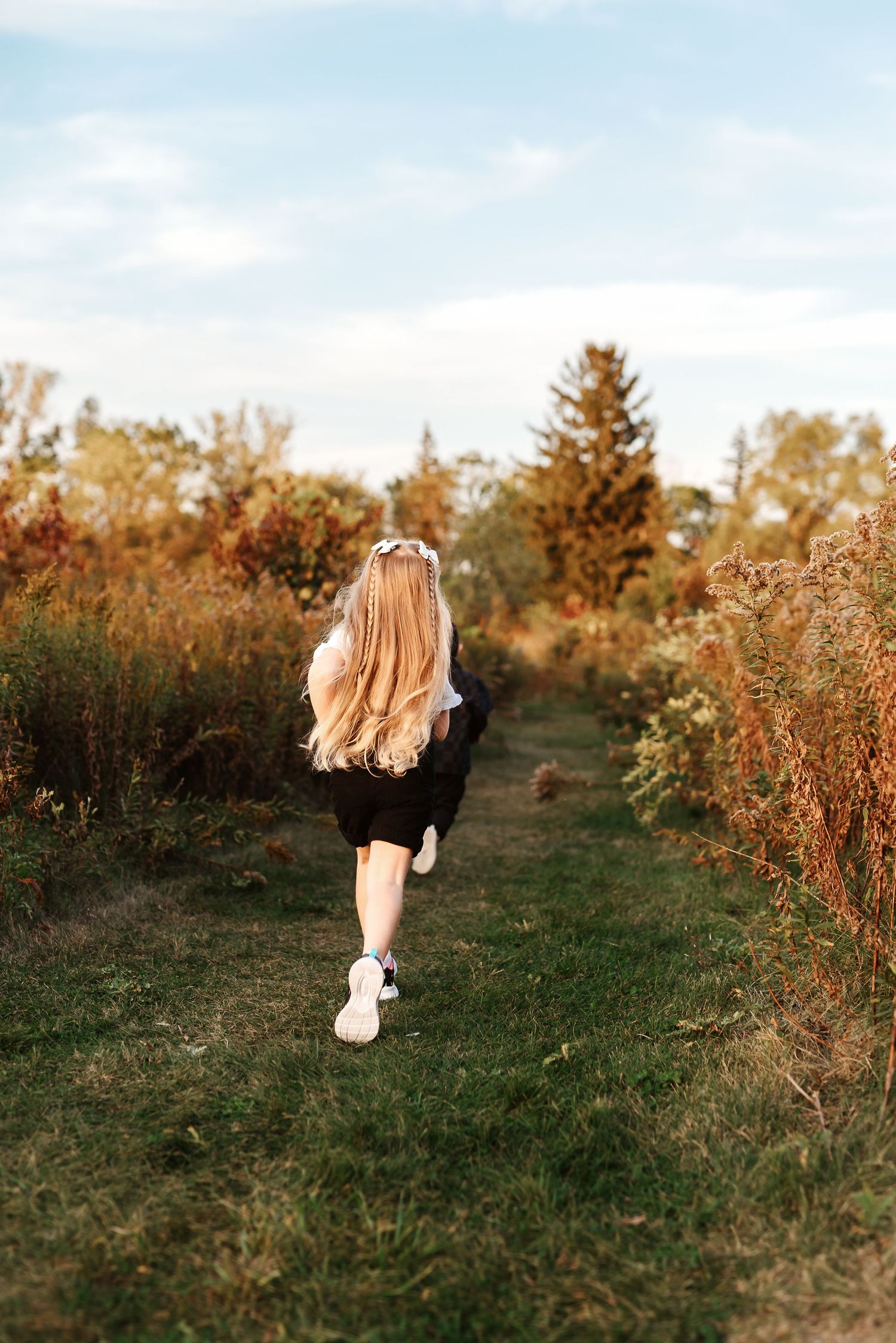 A person with blonde hair runs through a golden field at sunset with tall grass and trees in the background.