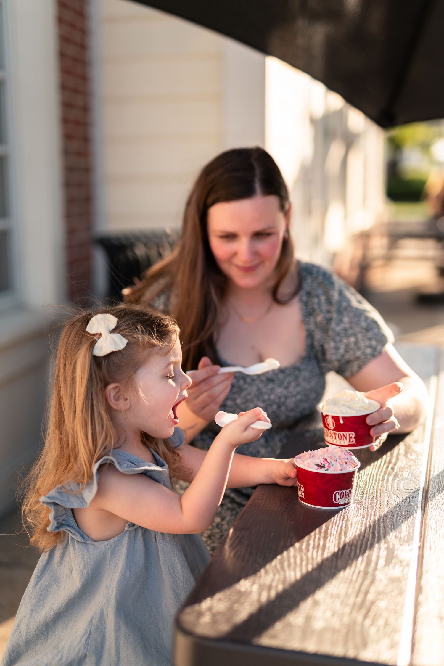A mother and young blonde girl share ice cream together at an outdoor wooden table in warm golden sunlight.