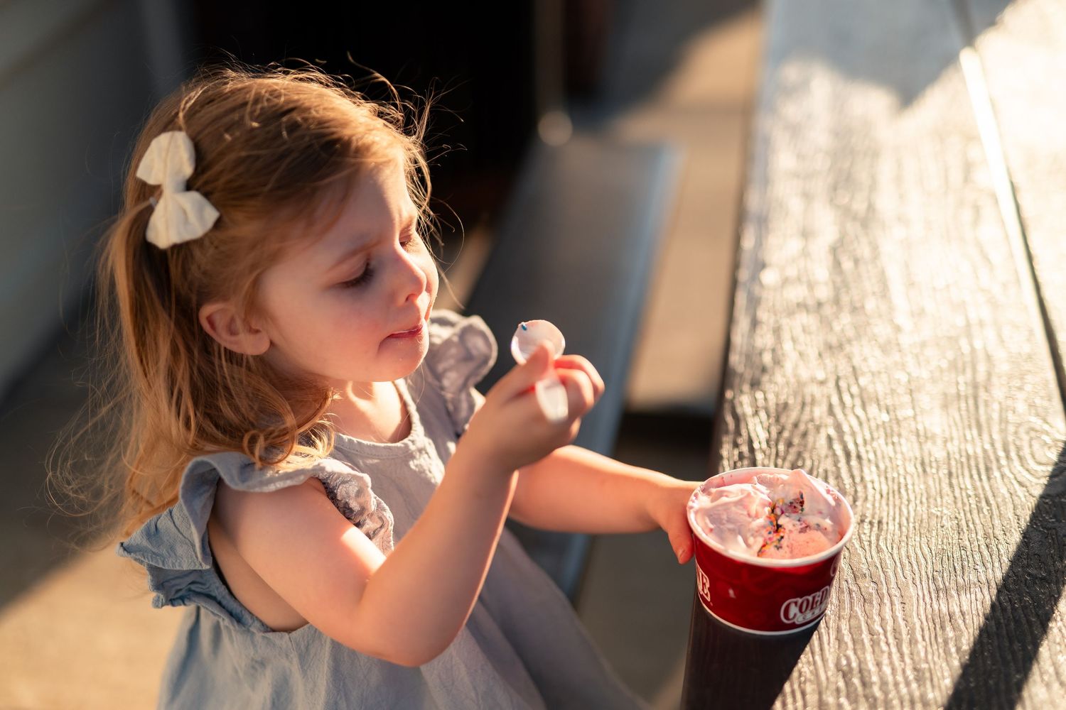 Young girl in blue dress enjoying a red slushy drink outdoors on a sunny day.