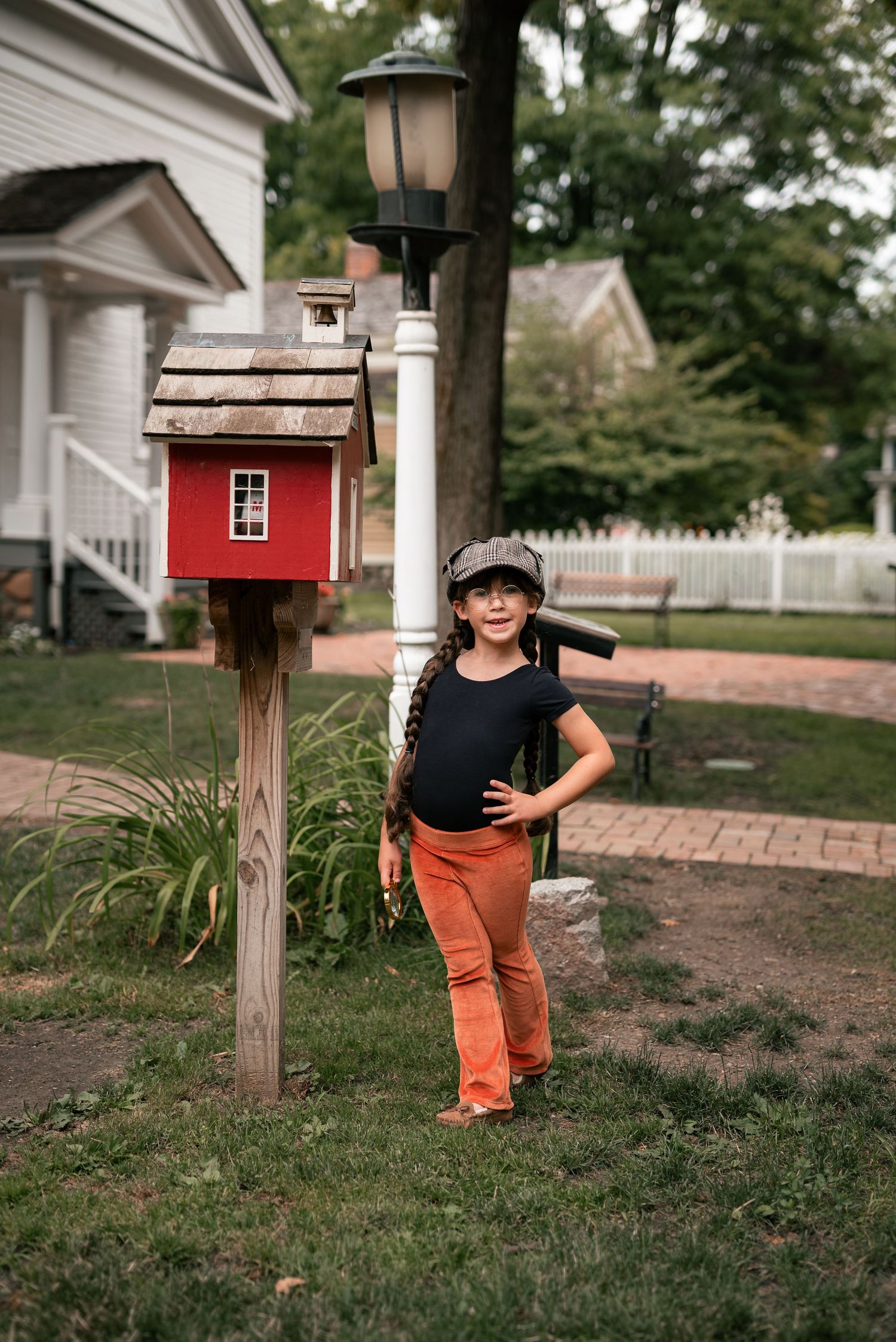 Young girl in orange pants and cap poses beside red miniature birdhouse on white lamppost.