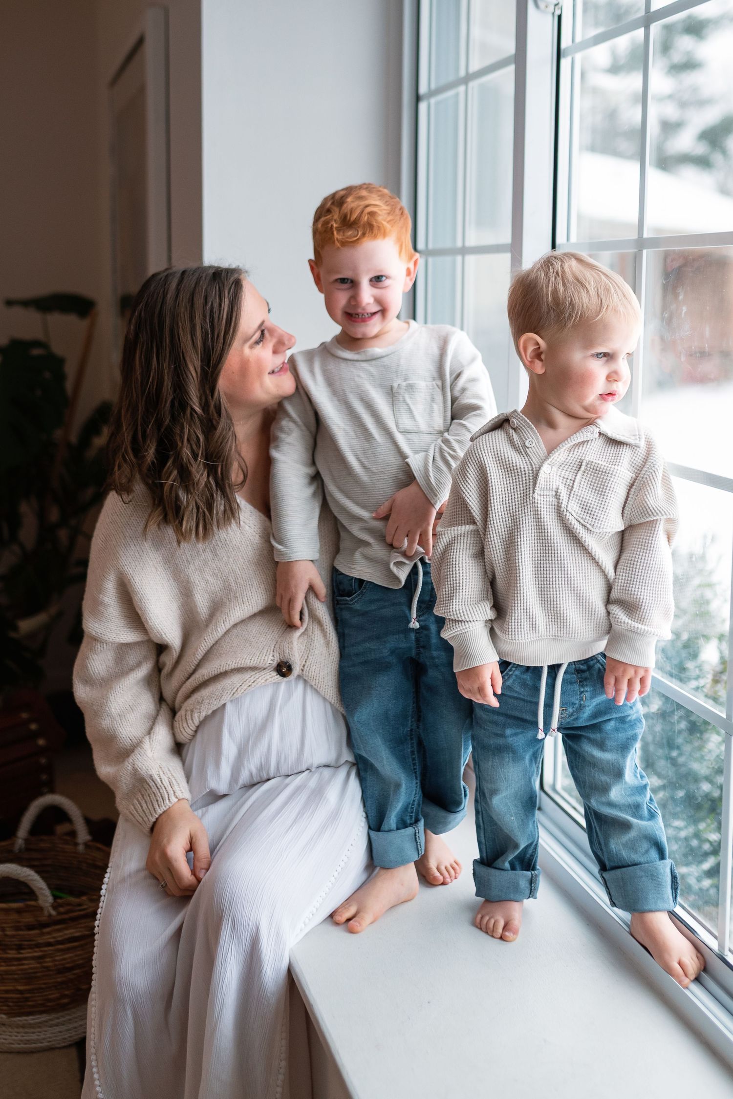 A mother sits on a windowsill smiling at her two young red and blonde-haired boys in cozy neutral sweaters and jeans.