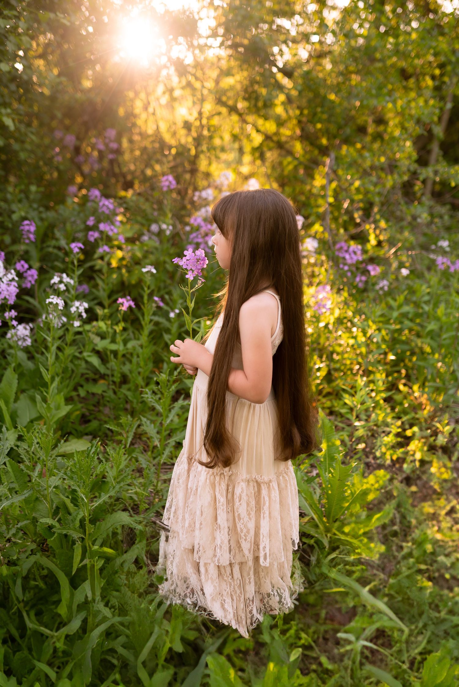 A person in a cream-colored dress stands among purple wildflowers during golden hour in a natural outdoor setting.