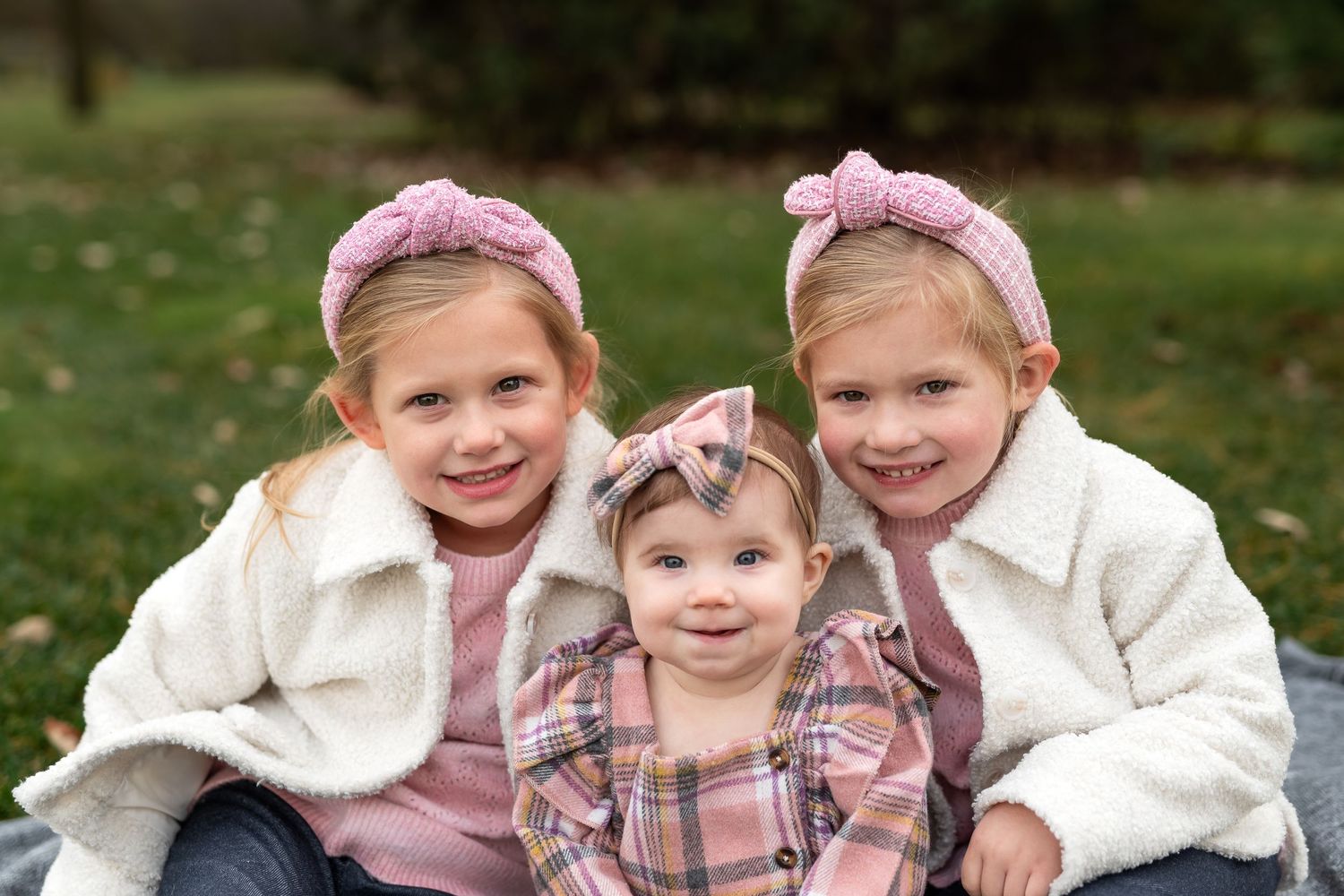 Three sisters in white jackets and pink accessories sitting together outdoors.