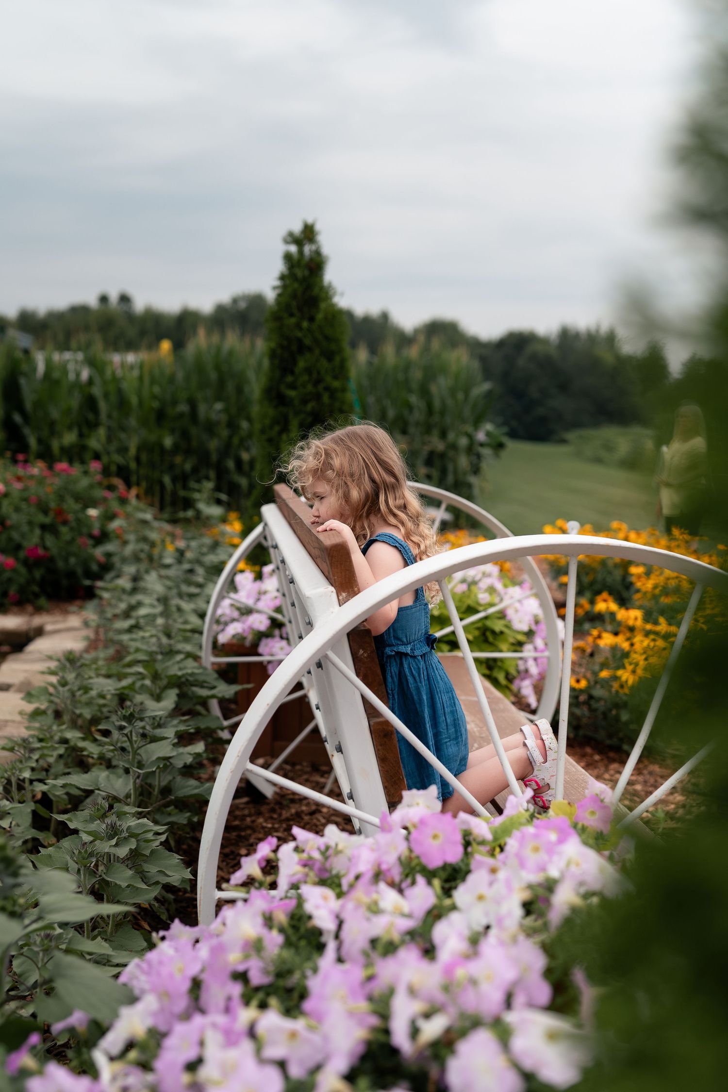 Young girl in blue dress sitting on white bench surrounded by colorful garden flowers.