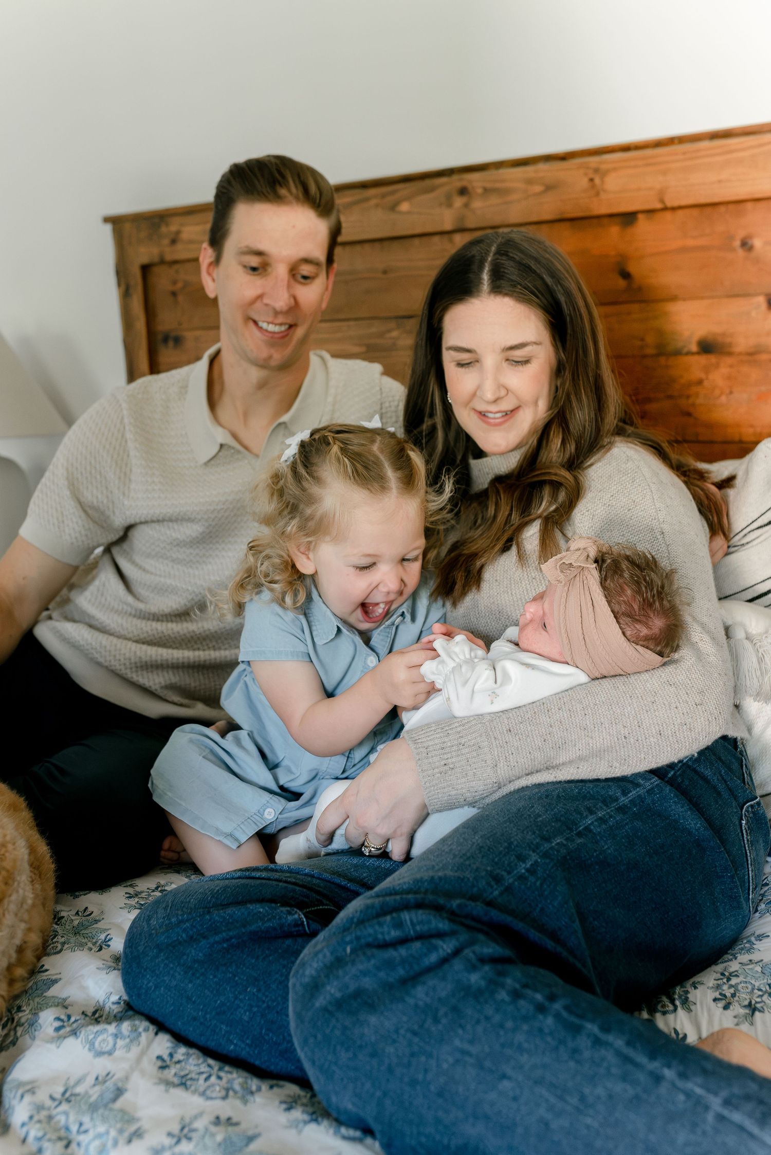 Happy family of four with toddler girl and newborn baby sitting together on bed.