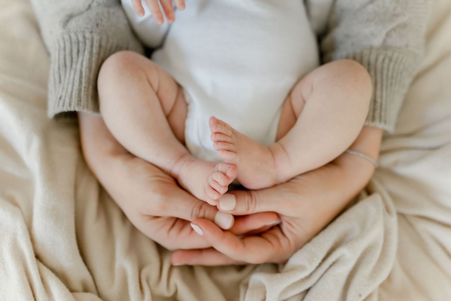 Close-up of a newborn baby's tiny feet cradled gently in loving parent's hands on a soft beige blanket.