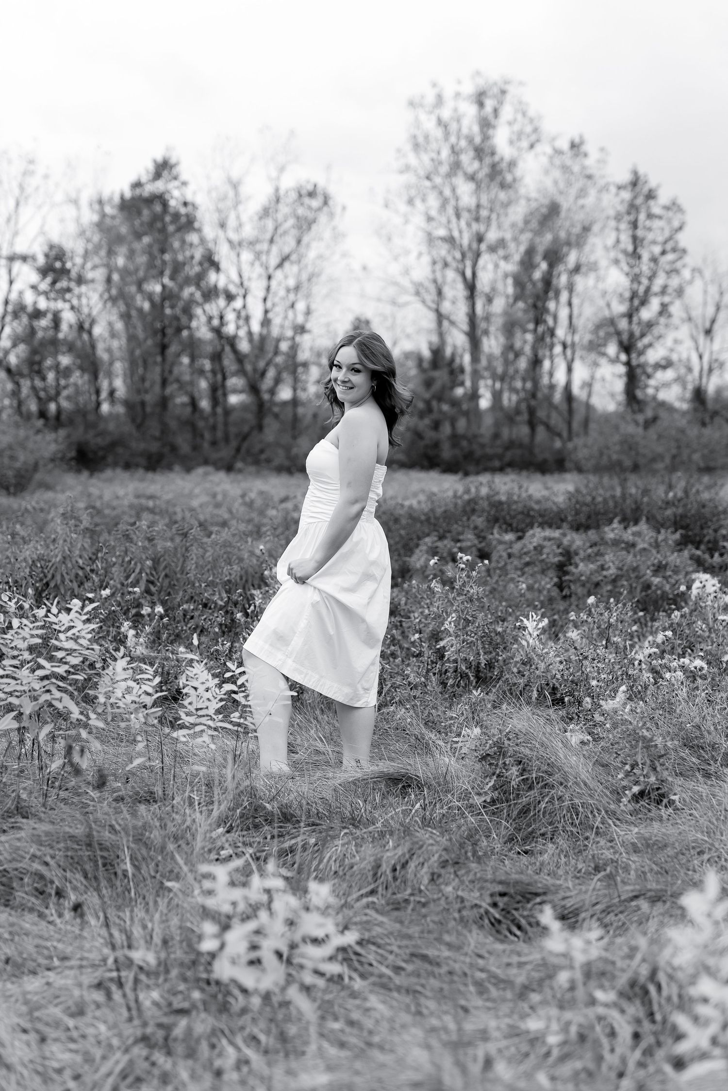 Woman in white dress posing in a wildflower field with bare trees in the background.
