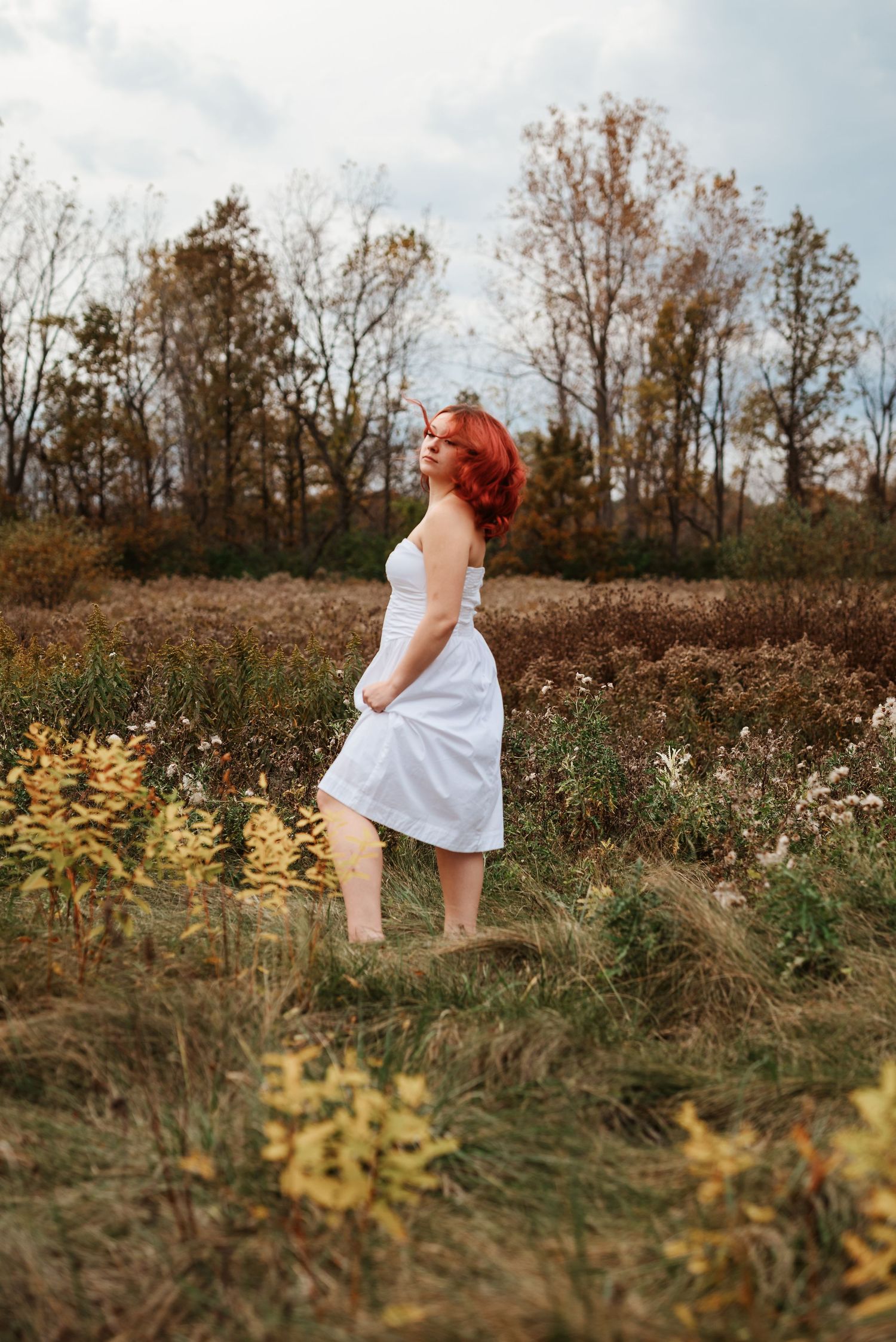 Red-haired woman in white dress standing in autumn field with colorful foliage and trees.