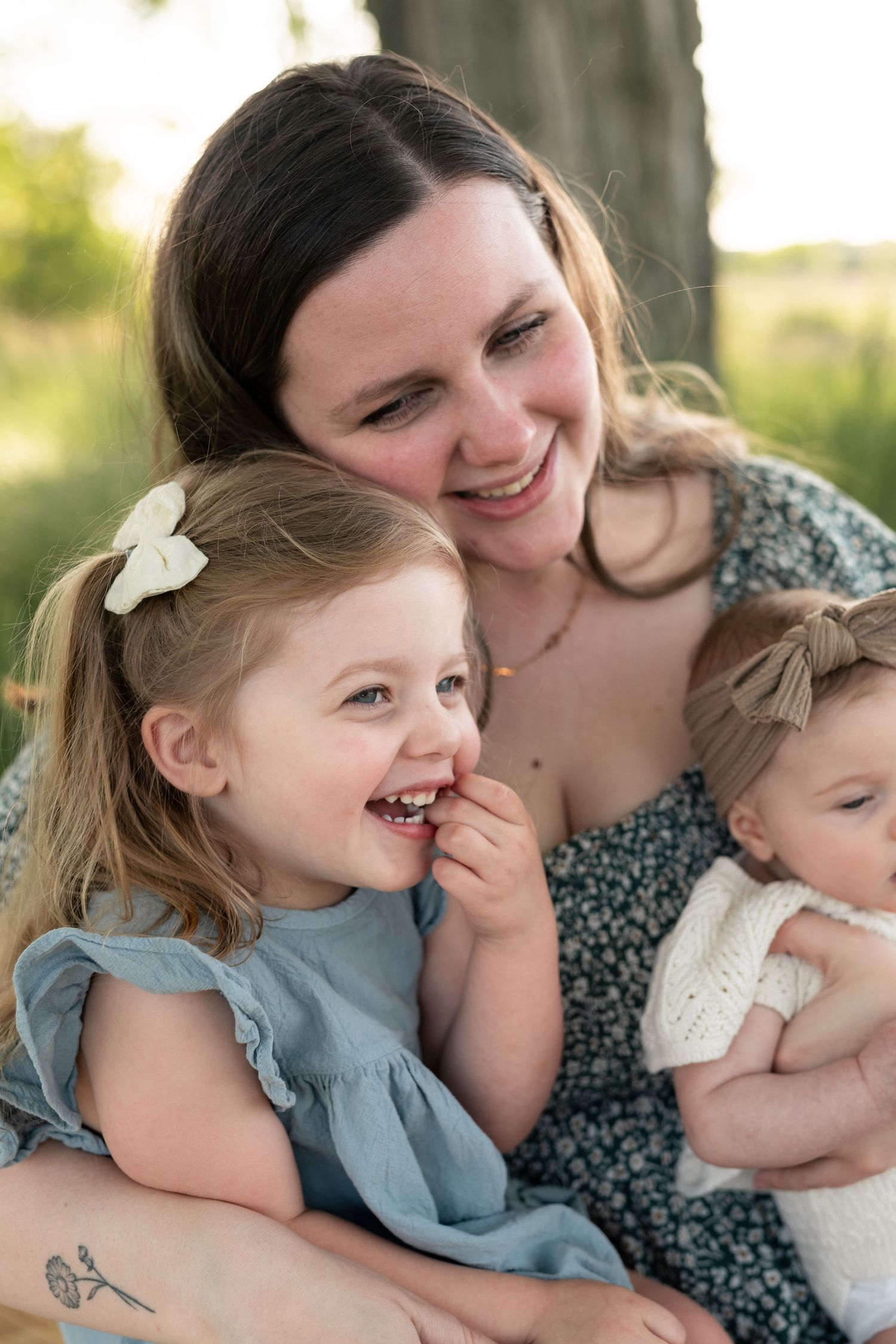 A smiling mother holds her newborn baby while her toddler daughter in a blue dress laughs outdoors.