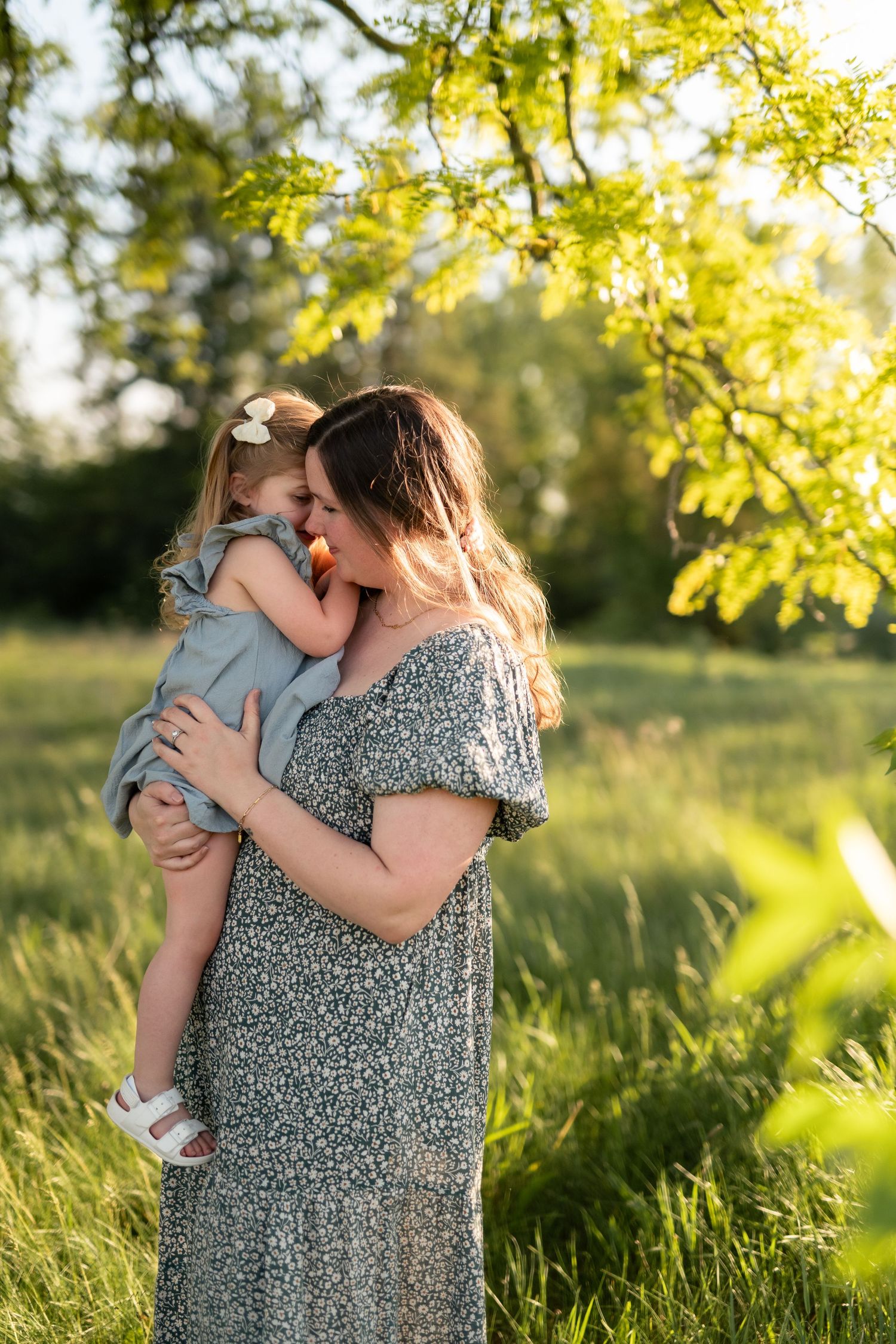 A mother holds and kisses her toddler daughter outdoors in golden sunlight, both wearing matching gray dresses.