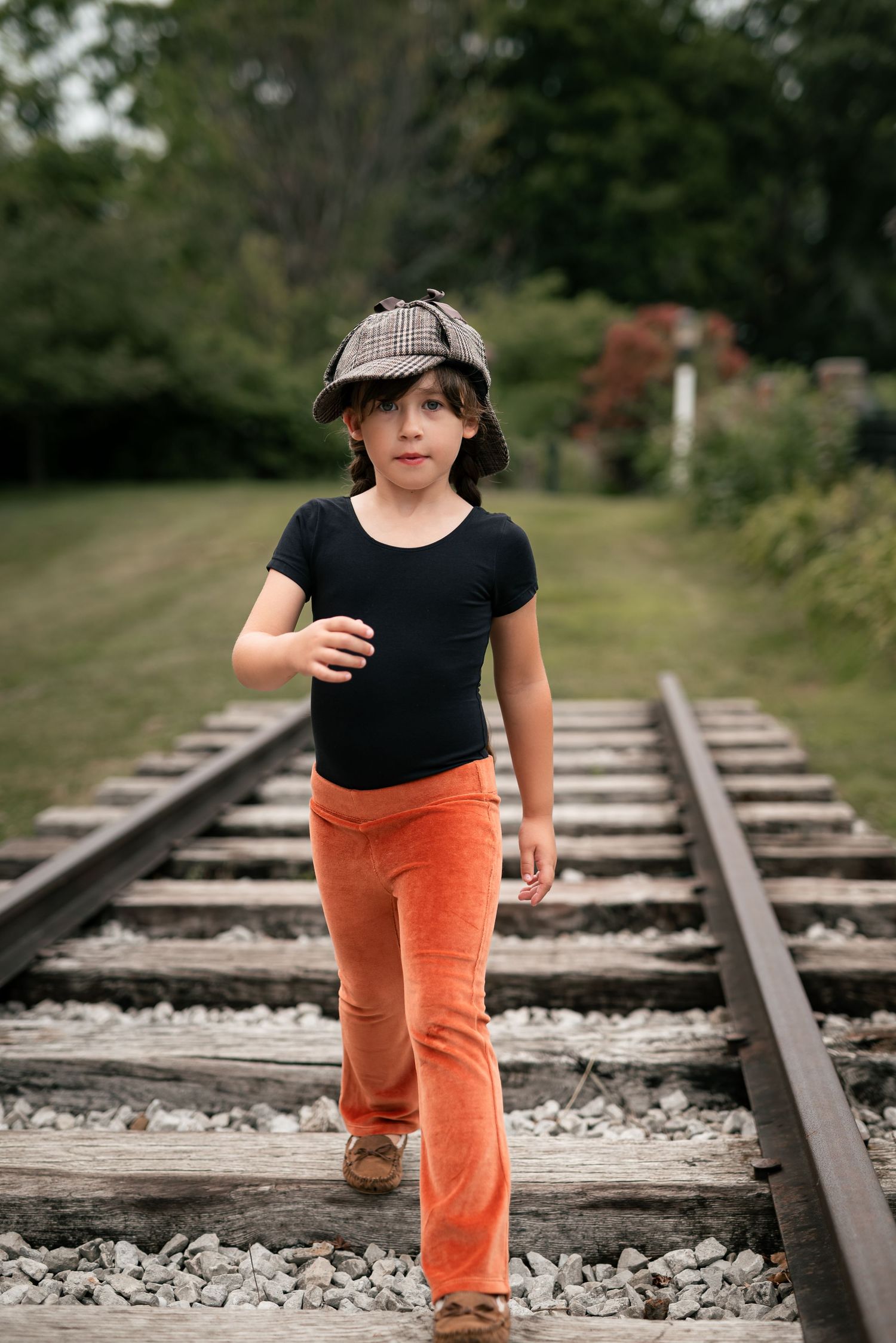 Child in orange pants and plaid hat walking on railroad tracks surrounded by green trees.