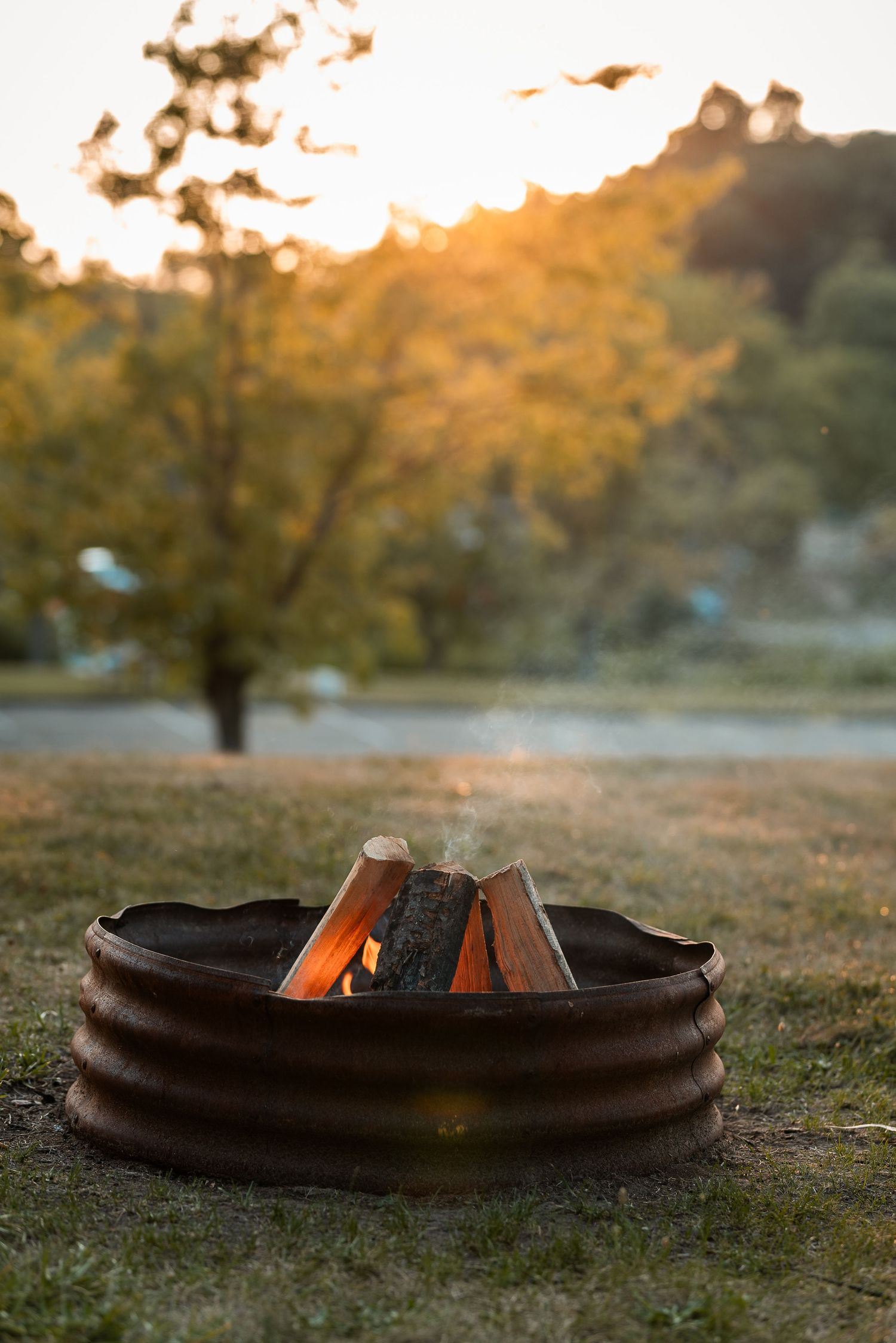 A rustic metal fire ring with burning logs glows warmly at sunset in a grassy campground with autumn trees in background.