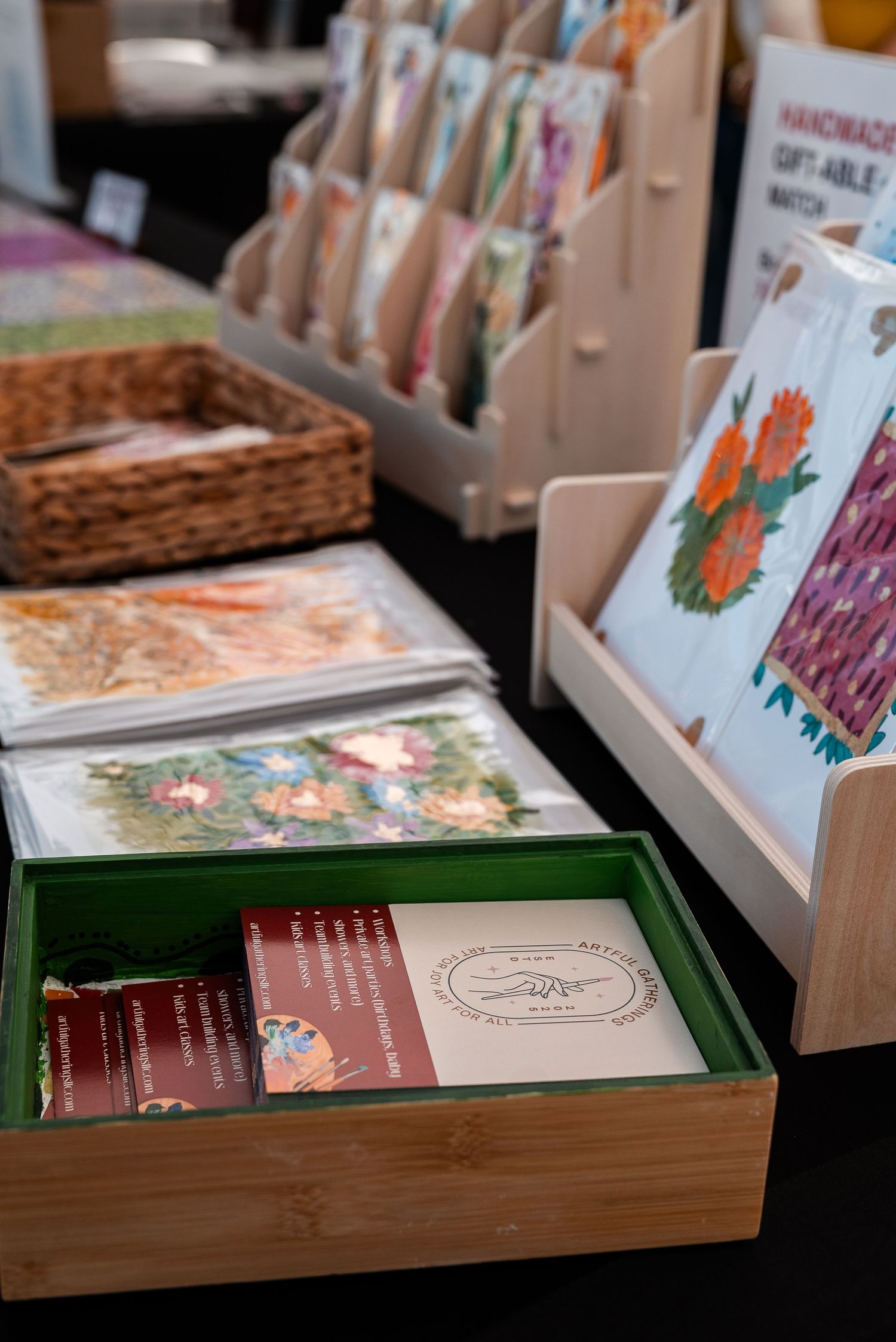 Handmade greeting cards and botanical art prints displayed on wooden stands at an indoor craft fair booth.