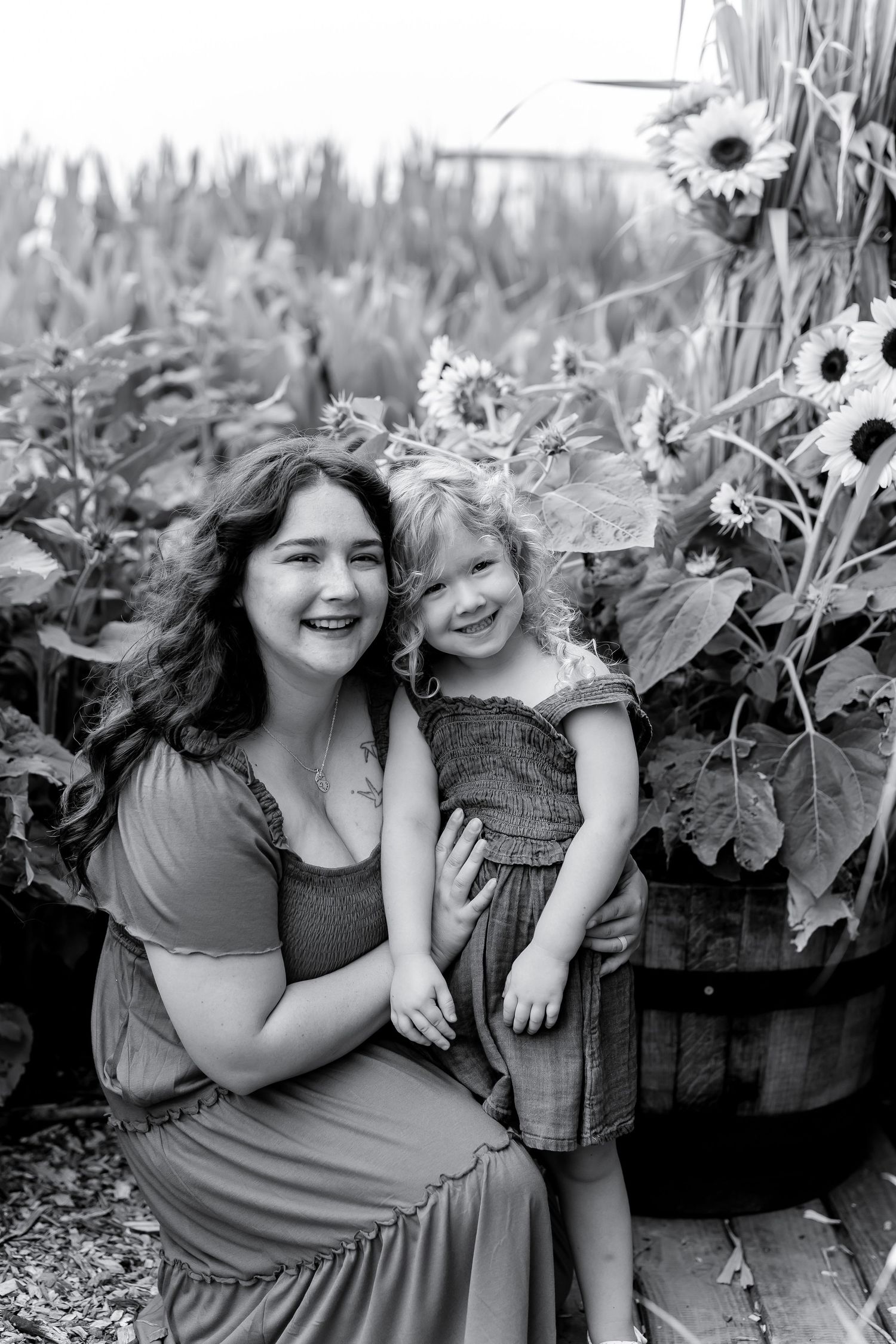 Black and white photo of a mother and young daughter smiling together among sunflowers and garden plants.