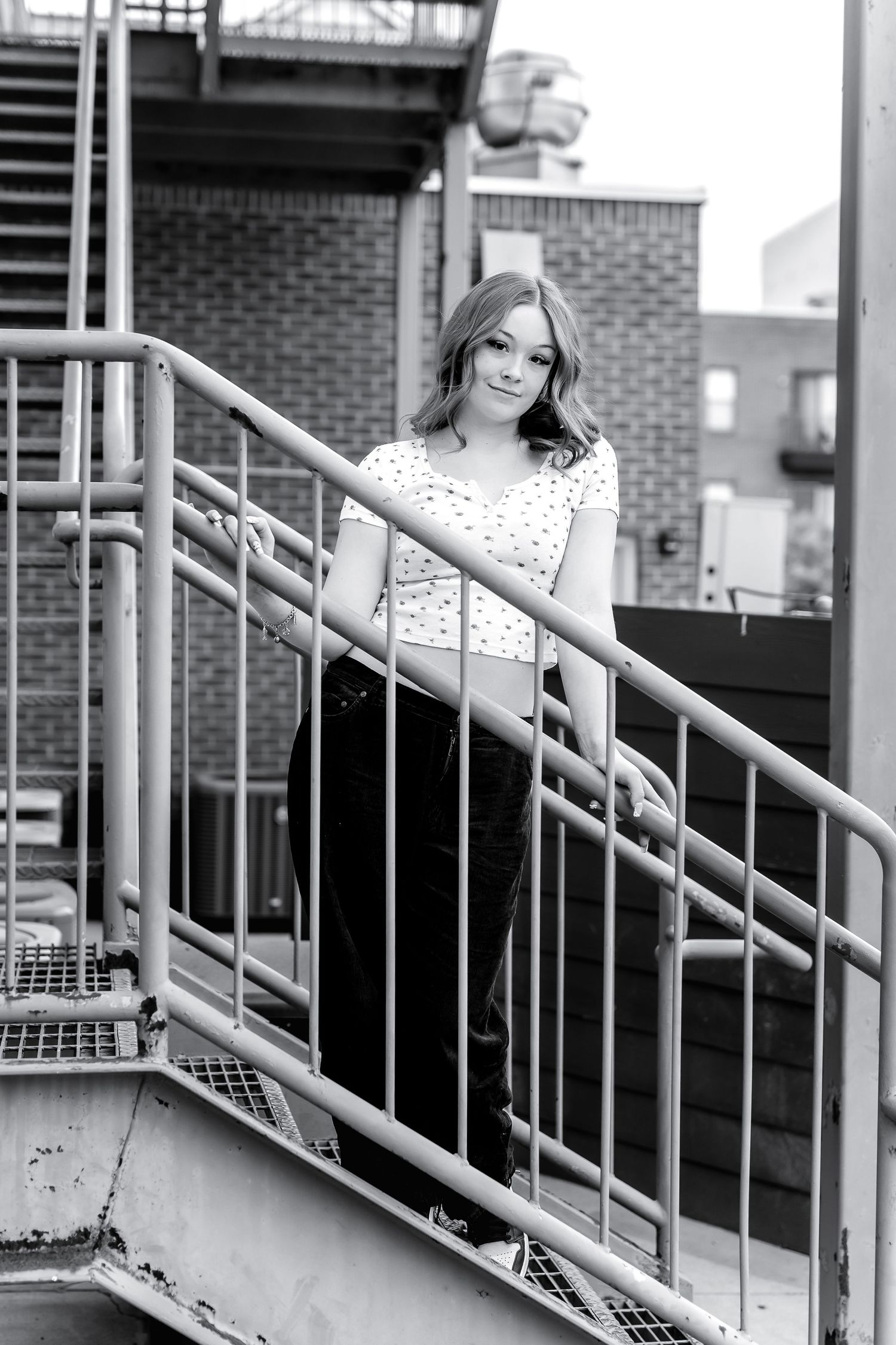 Black and white photo of young woman posing on metal fire escape staircase of brick building.