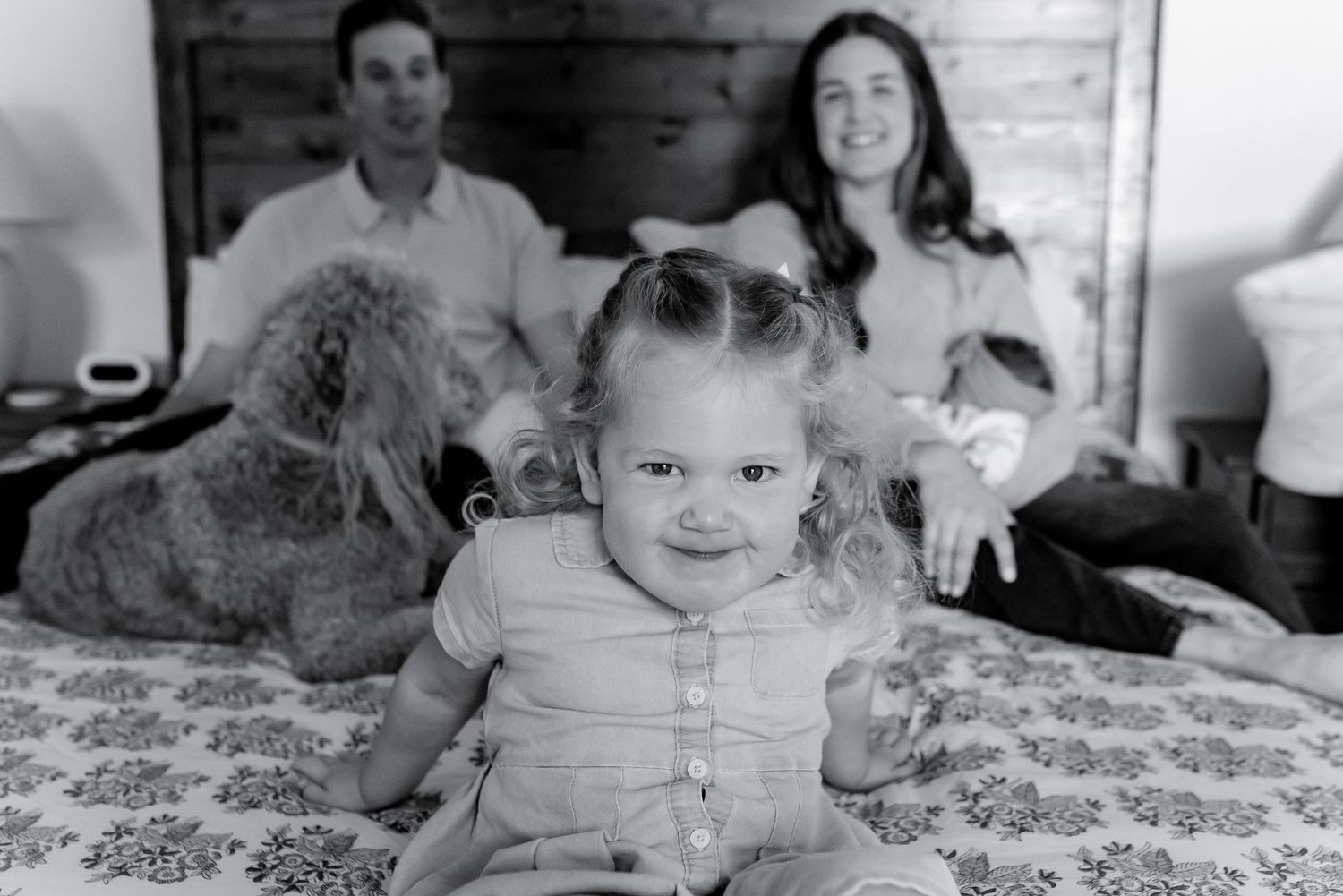 Black and white family photo with parents, toddler girl, and fluffy dog sitting together on a bed.