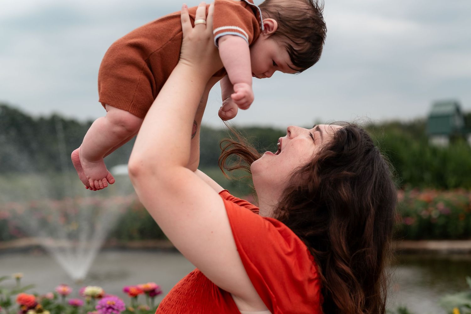 A joyful mother lifts her smiling baby in the air outdoors near a fountain and colorful flowers.