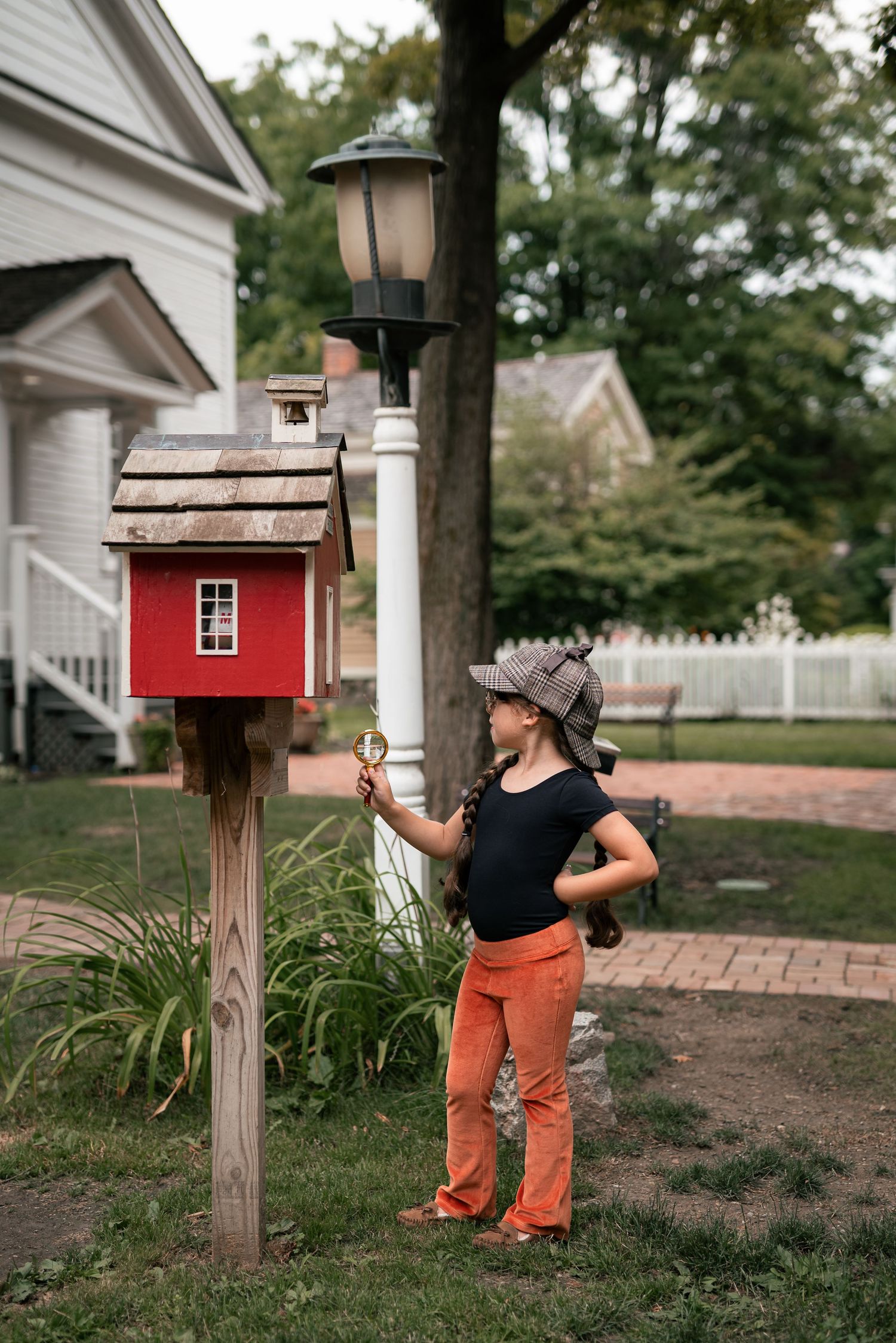 Girl in detective hat holds magnifying glass near red birdhouse on post in suburban backyard.