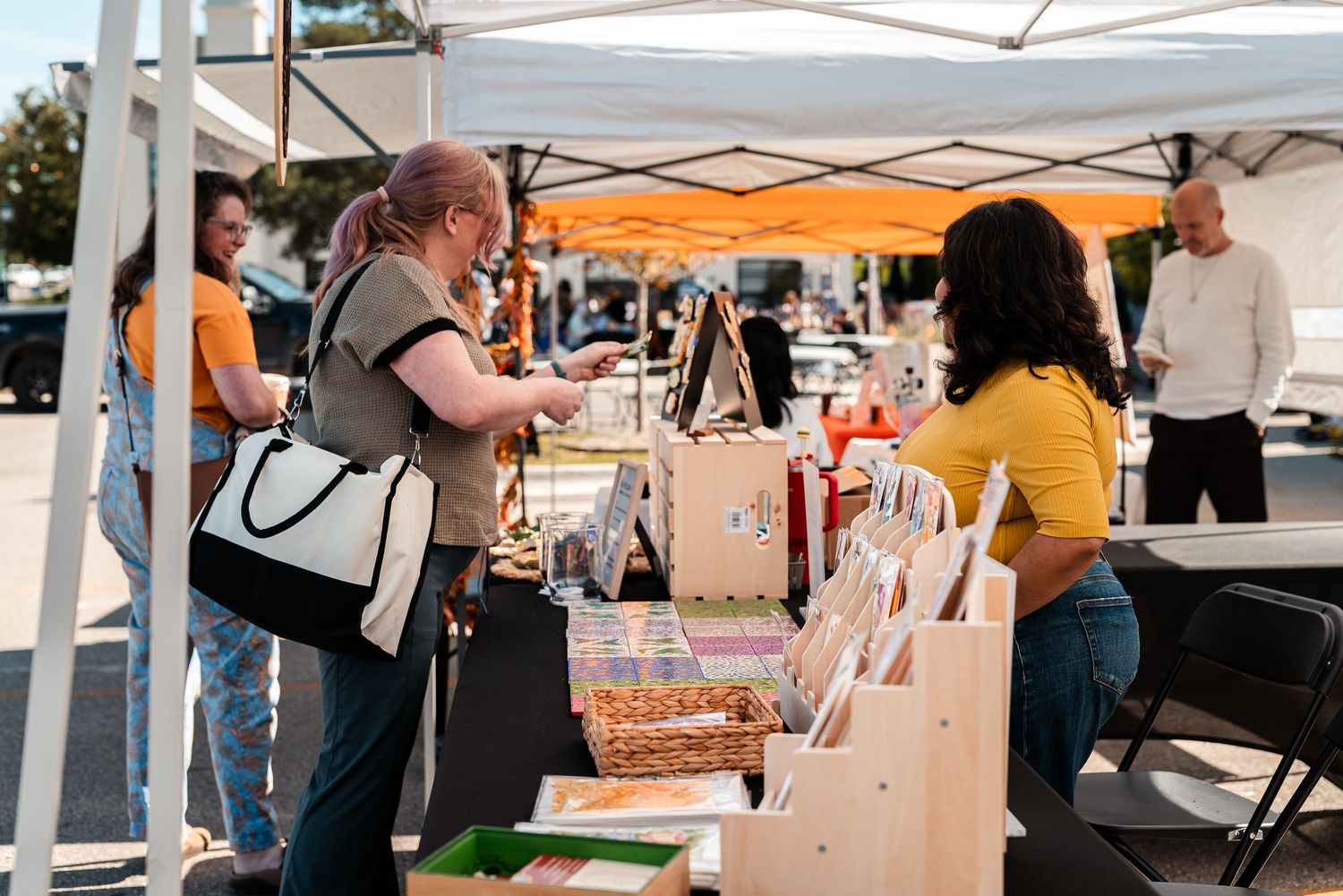 Shoppers browsing handmade goods at an outdoor market booth under a white canopy tent on a sunny day.