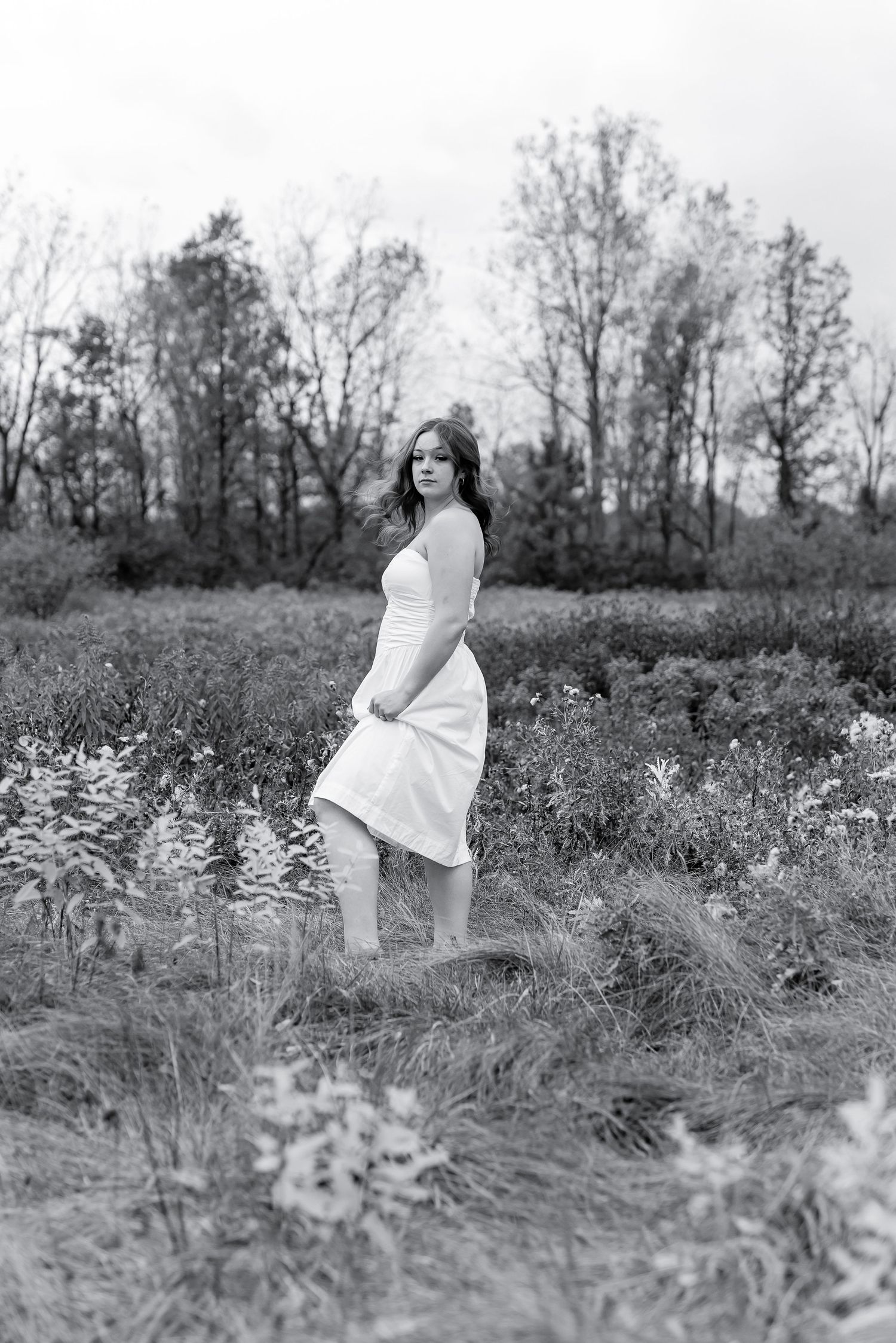 Young woman in flowing white dress posing in overgrown field in black and white.