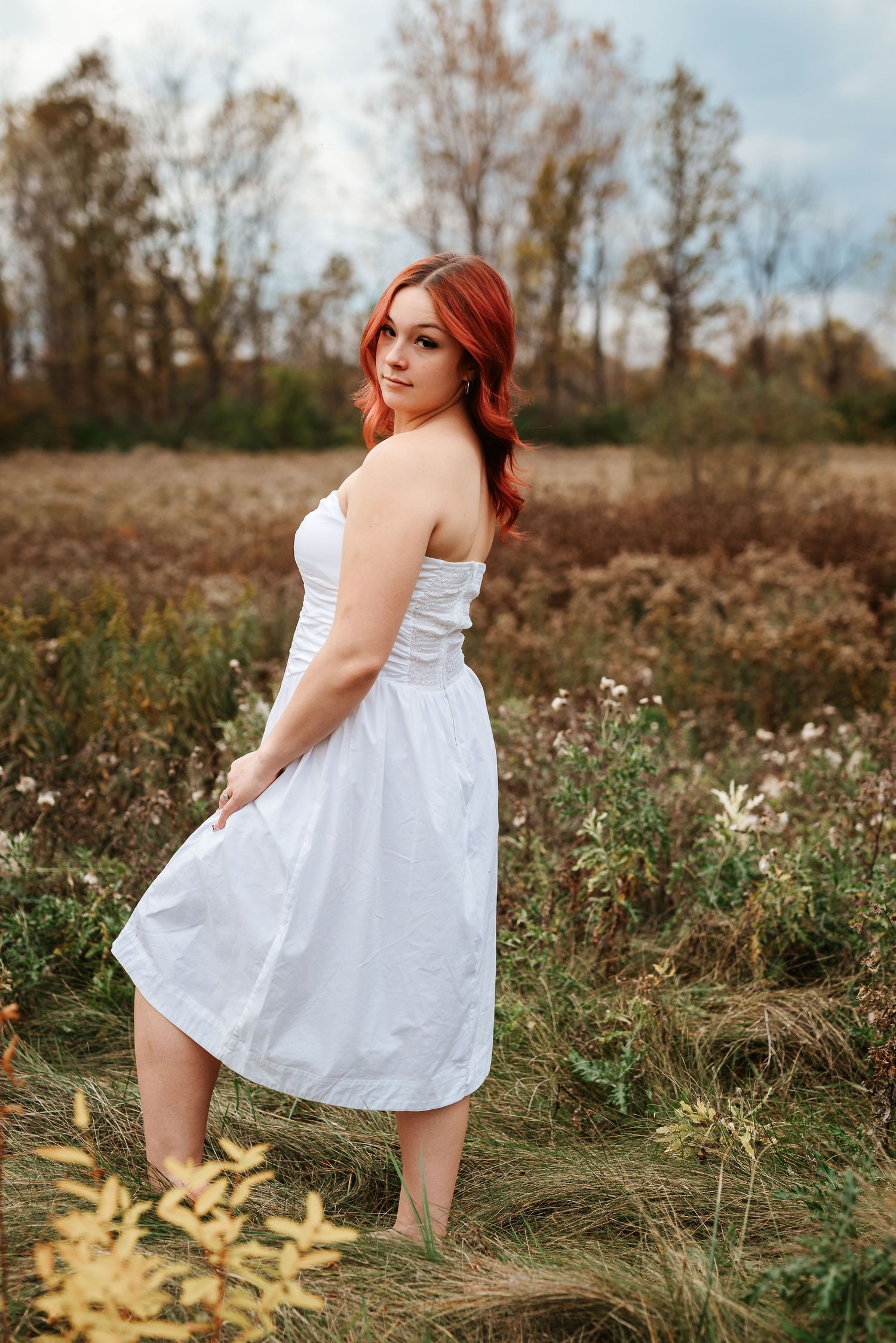 Young woman with red hair wearing a white strapless dress posing in a fall meadow landscape.