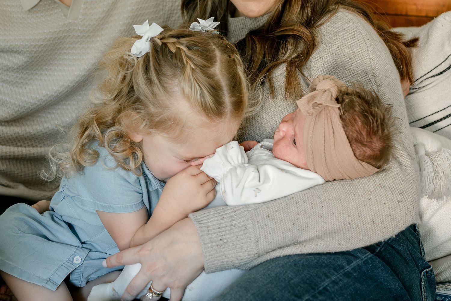 Toddler girl with curly hair kissing newborn baby sibling held by mother in cozy family photo.