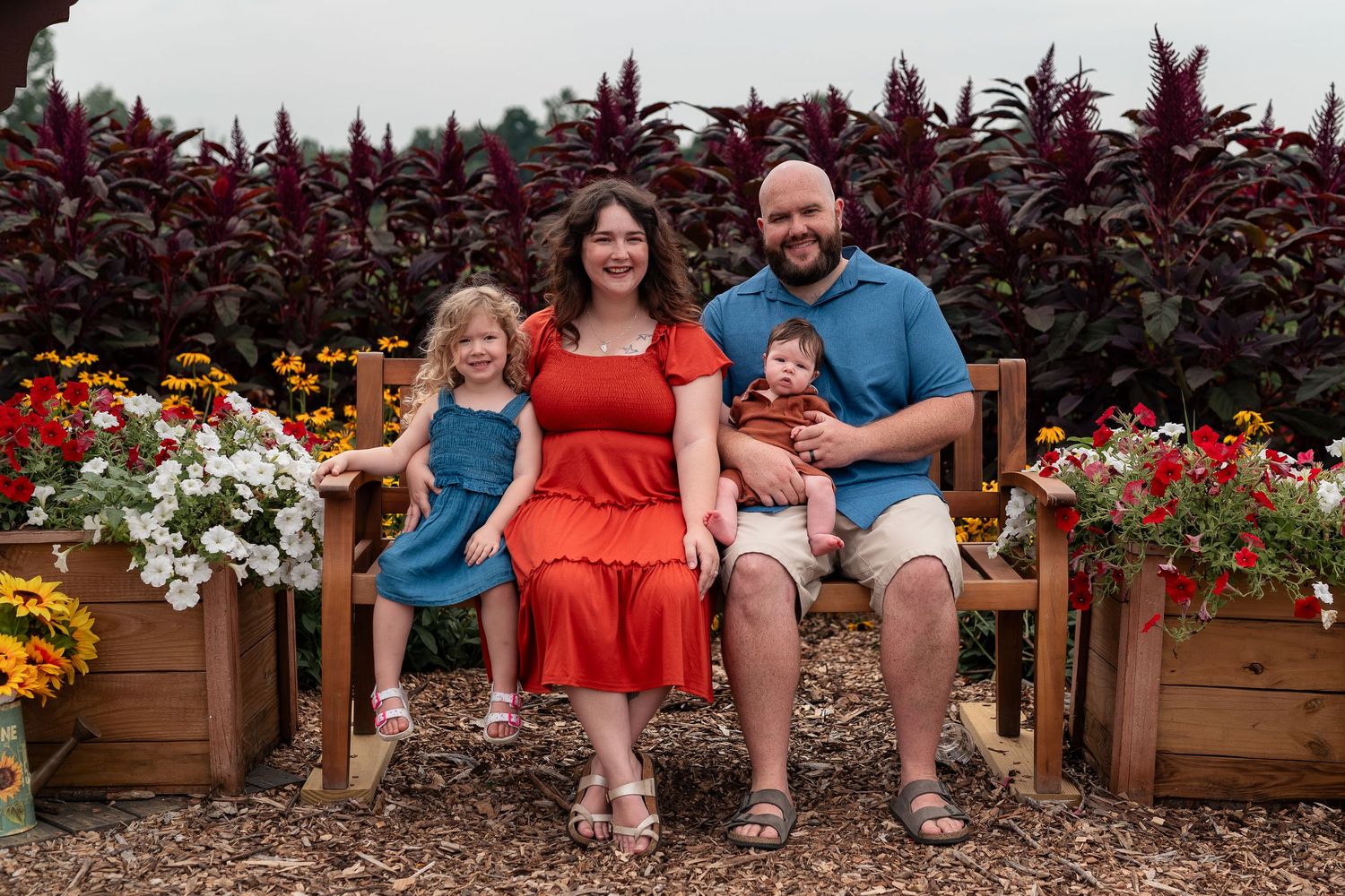 Family of four posing on a wooden bench surrounded by colorful flowers in a garden setting.