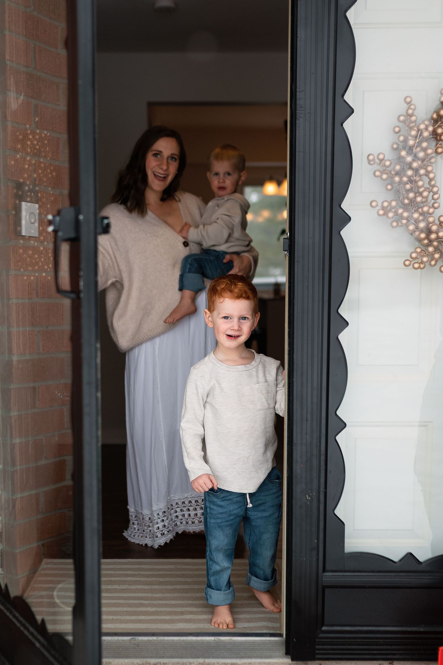 A smiling woman holding a toddler stands at an open front door with a redheaded boy in jeans standing in the doorway.