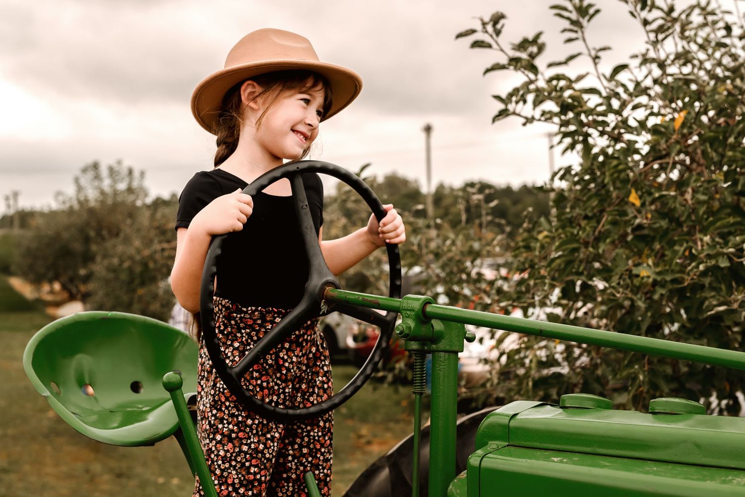Person in a polka dot dress and brown hat sits on a vintage green tractor in an orchard setting.