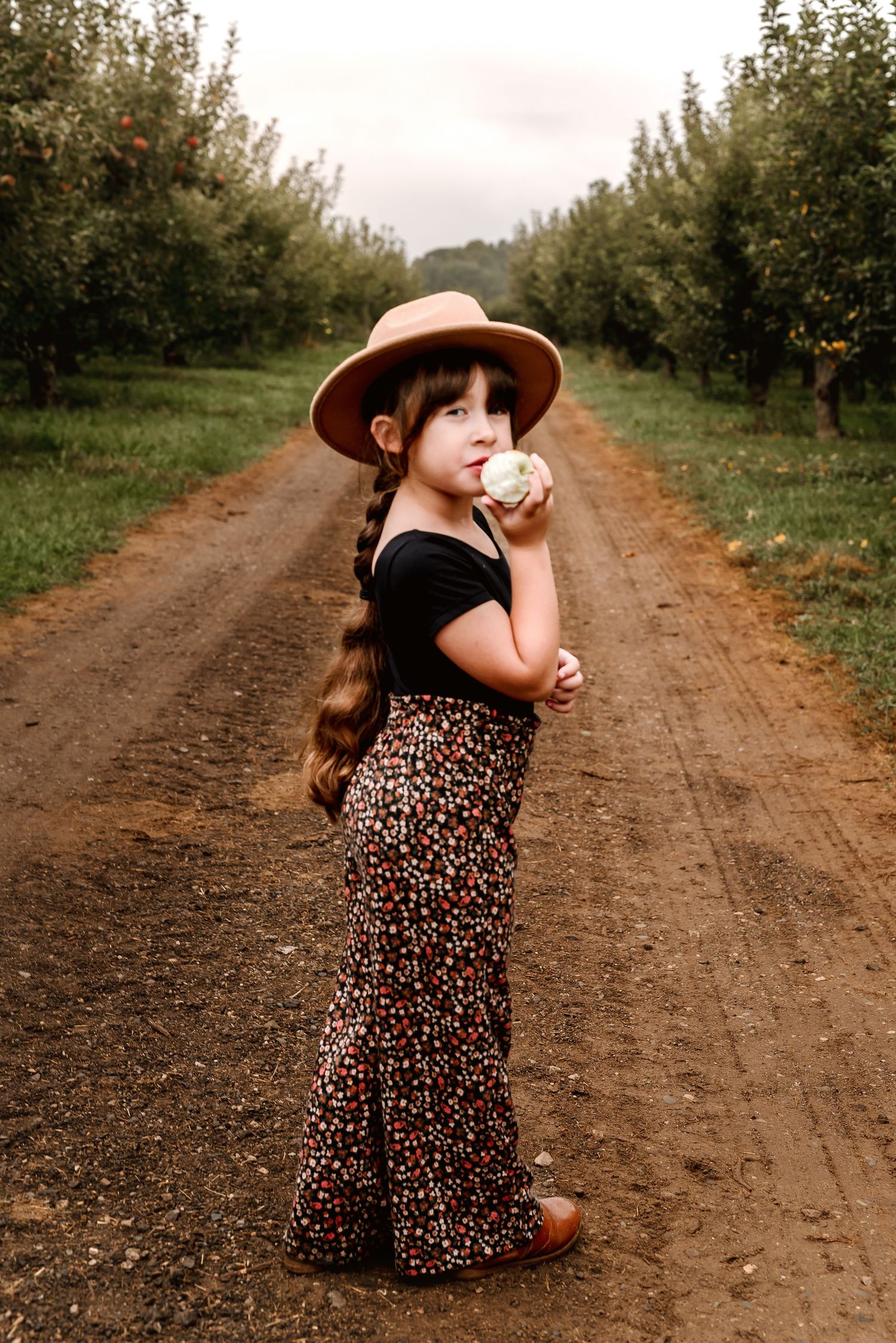 Child in a wide-brimmed hat and floral maxi skirt stands on a dirt path between rows of apple trees in an orchard.