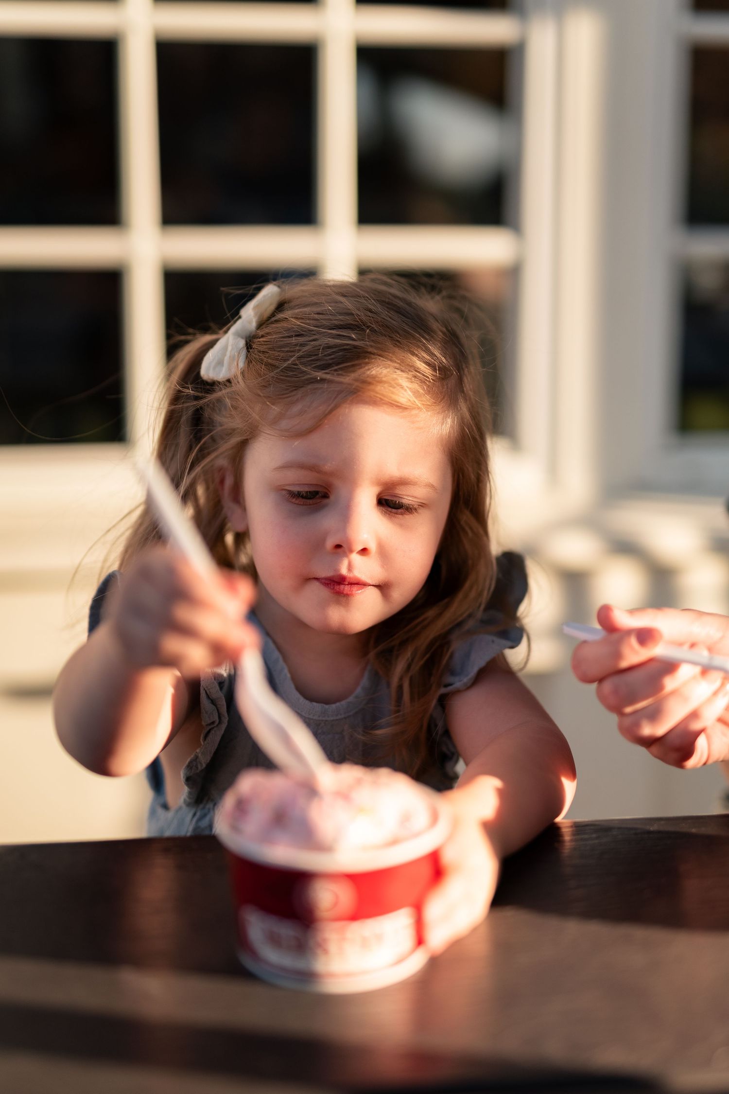 Young girl enjoying ice cream from a red cup at an outdoor table in warm golden sunlight.