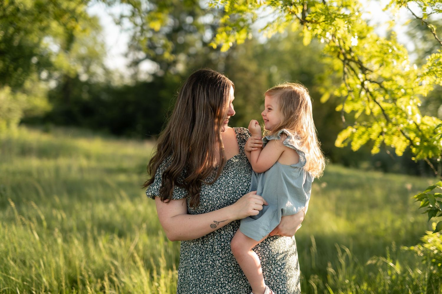 A mother holds her toddler daughter in a sunlit green meadow, sharing a tender moment outdoors.