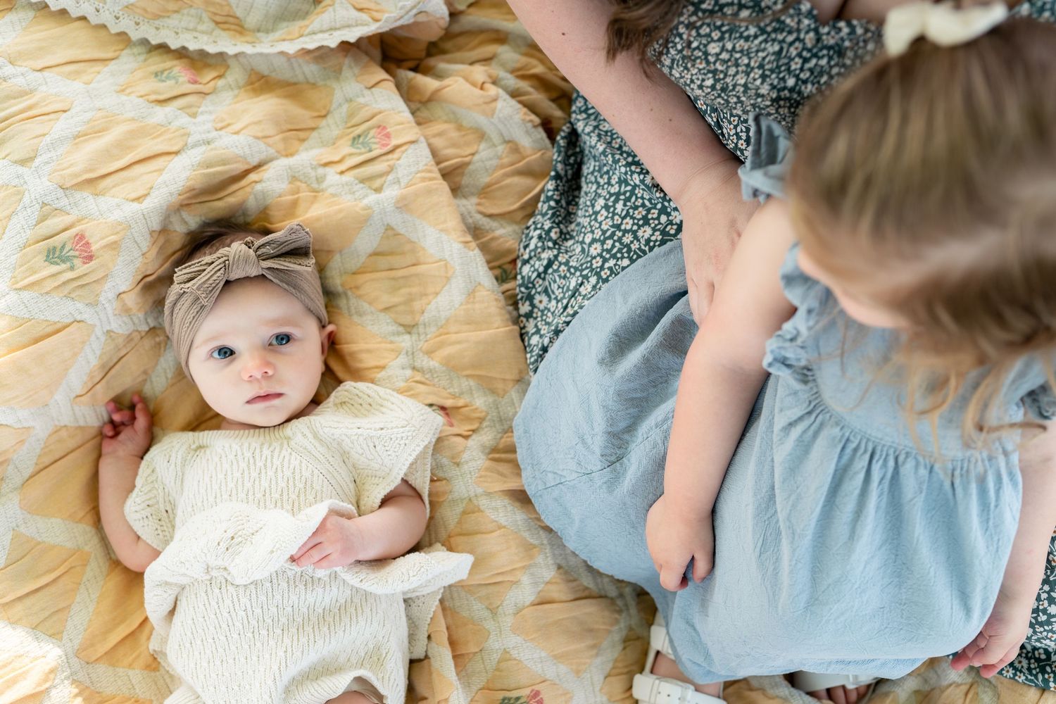 Baby girl in cream knit dress with headband lies on floral quilt beside older sister in blue dress.