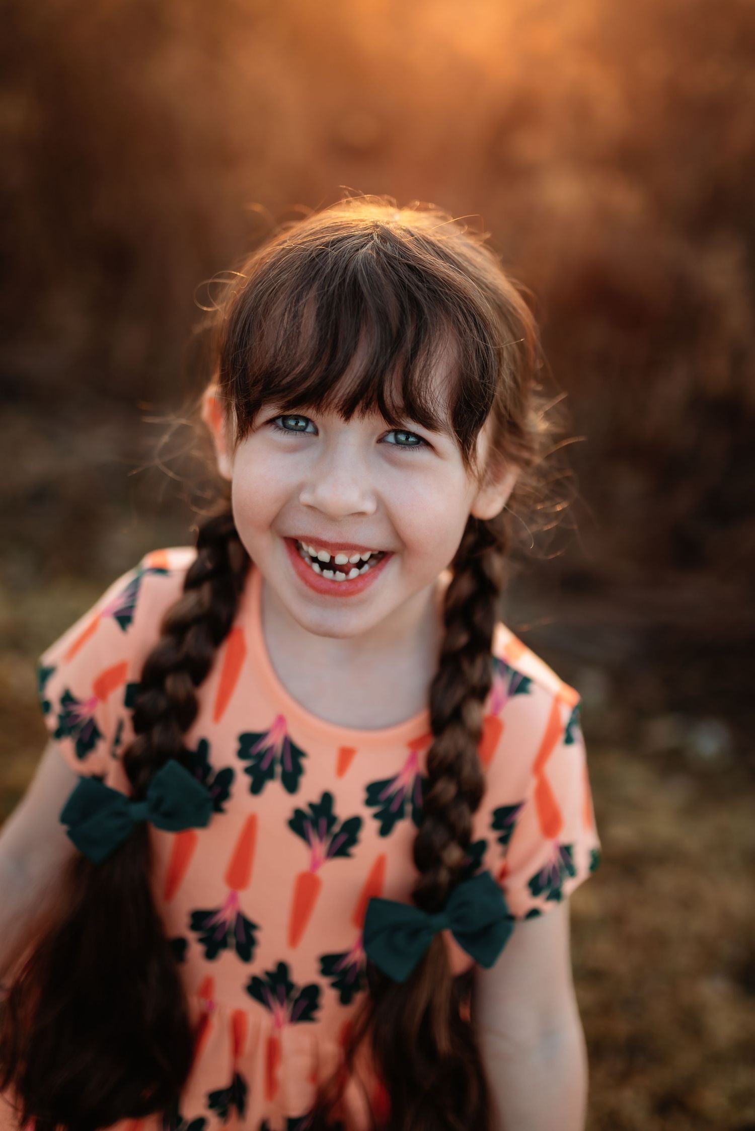 A young child in pigtails wearing a peach-colored floral dress smiles brightly during golden hour photography.