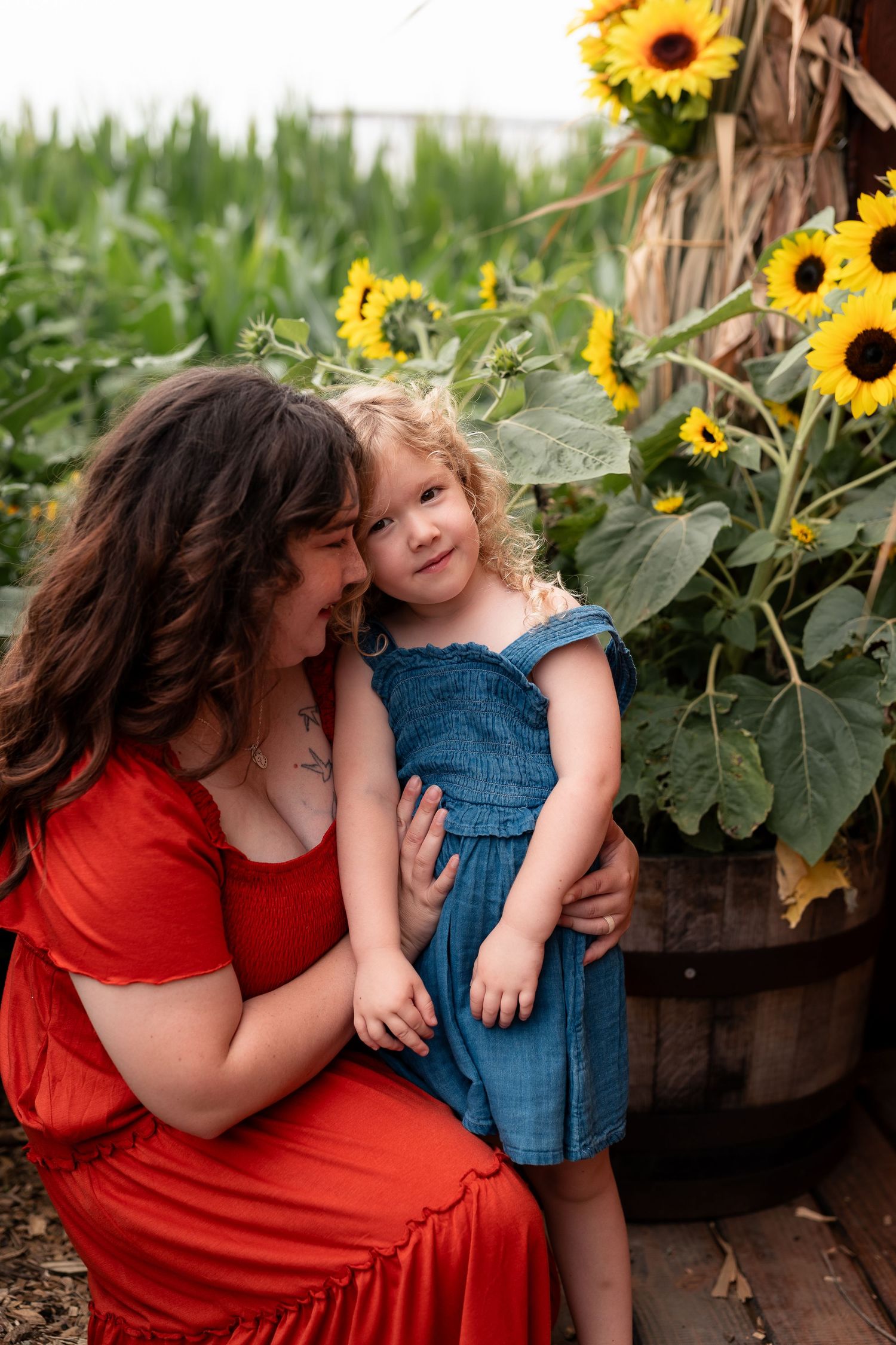 A woman in a red dress cuddles a curly-haired toddler girl in blue near vibrant sunflowers in a garden.