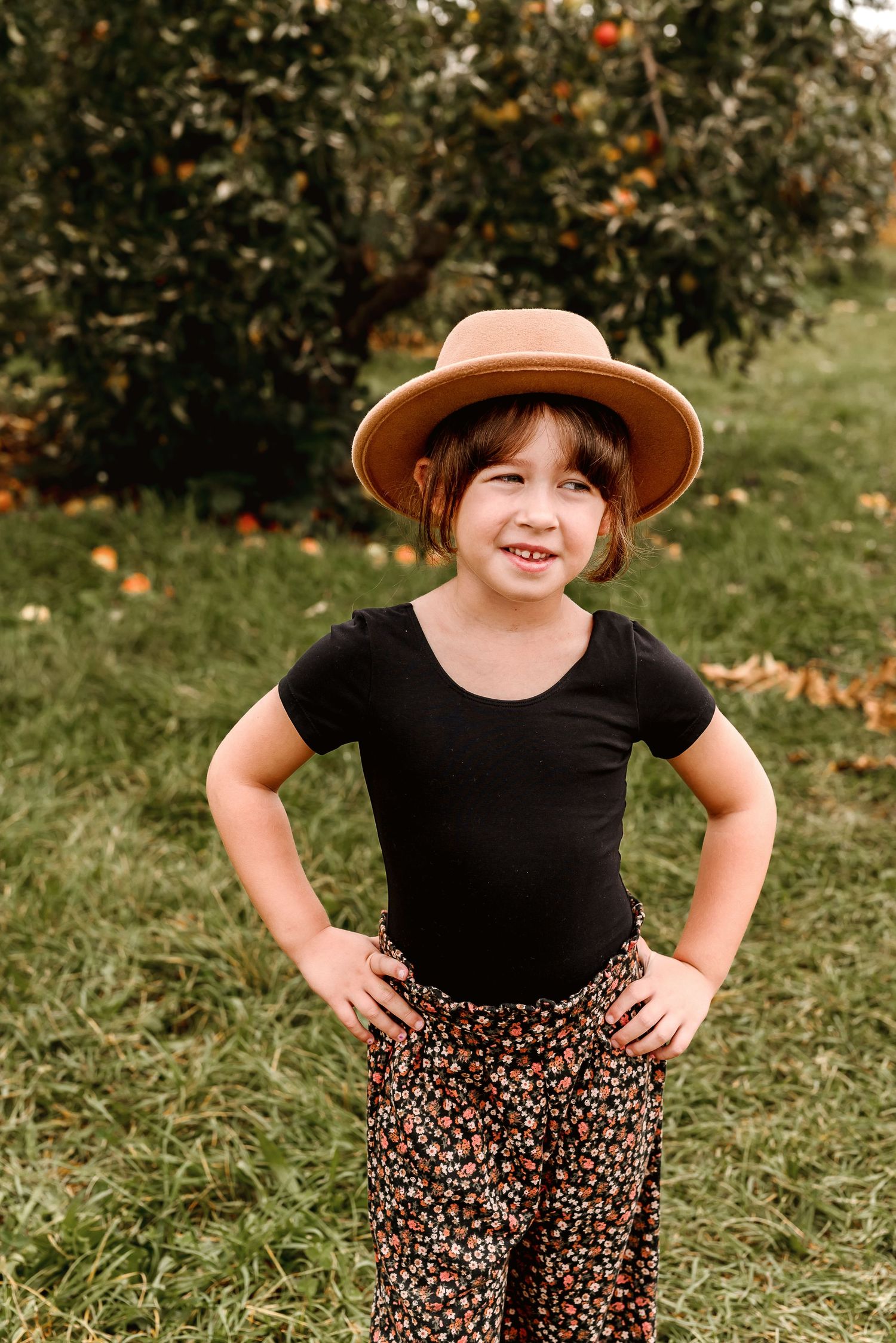 A child in a black top and floral pants stands playfully in a grassy outdoor setting with orange trees in background.