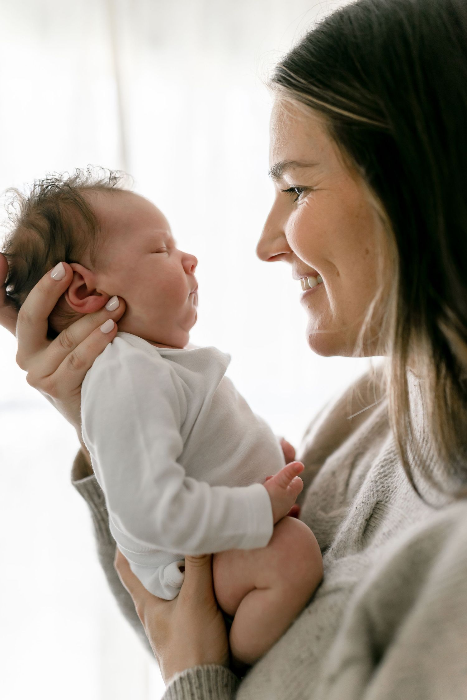Joyful mother in gray sweater smiling at sleeping newborn baby held in her arms.