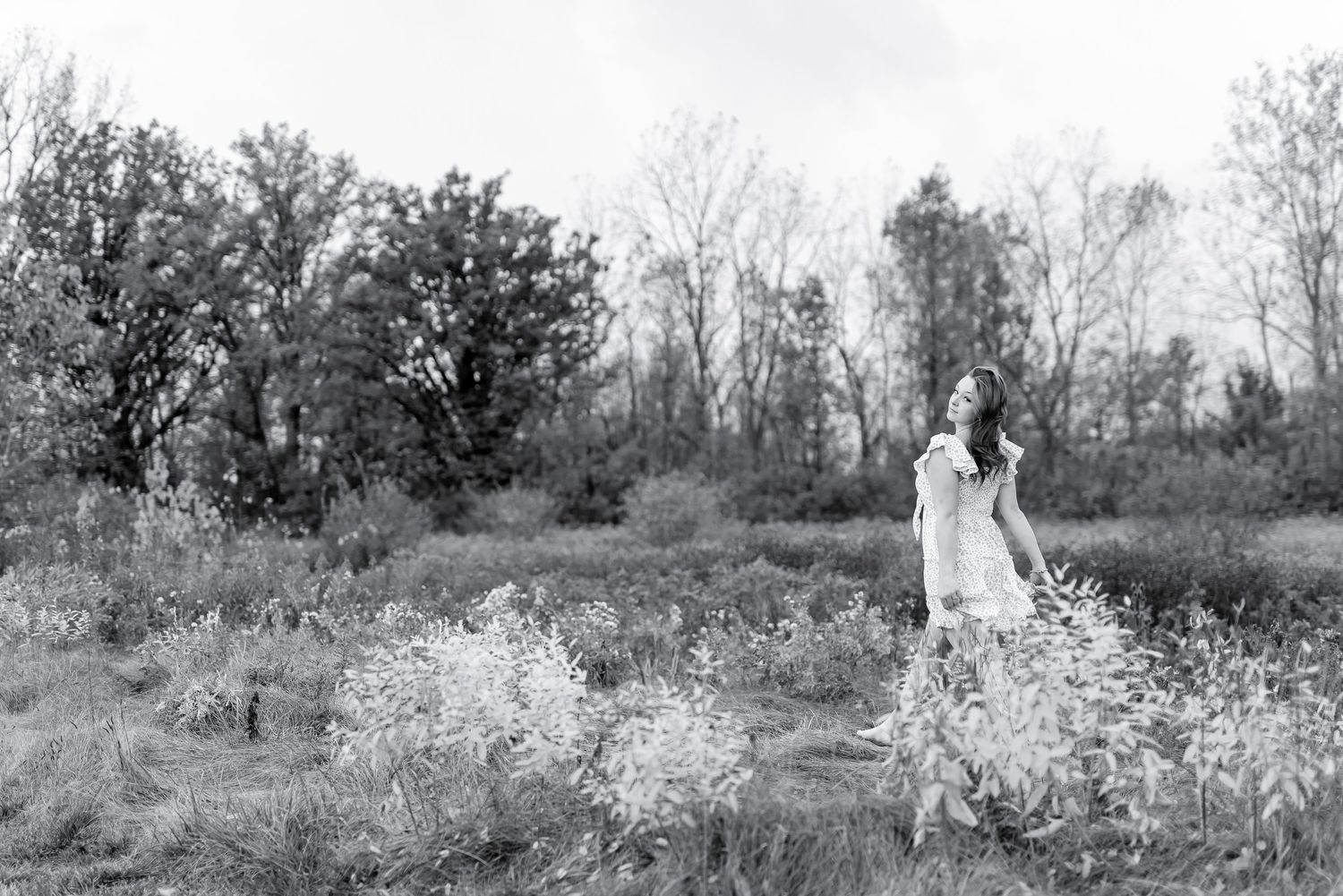 Black and white wide shot of young woman in white dress standing among wildflowers in open field.