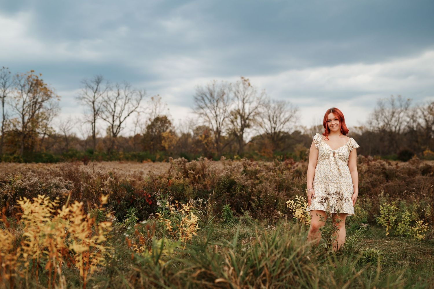 A young woman with red hair in a floral dress stands in an autumn field with bare trees under a cloudy sky.
