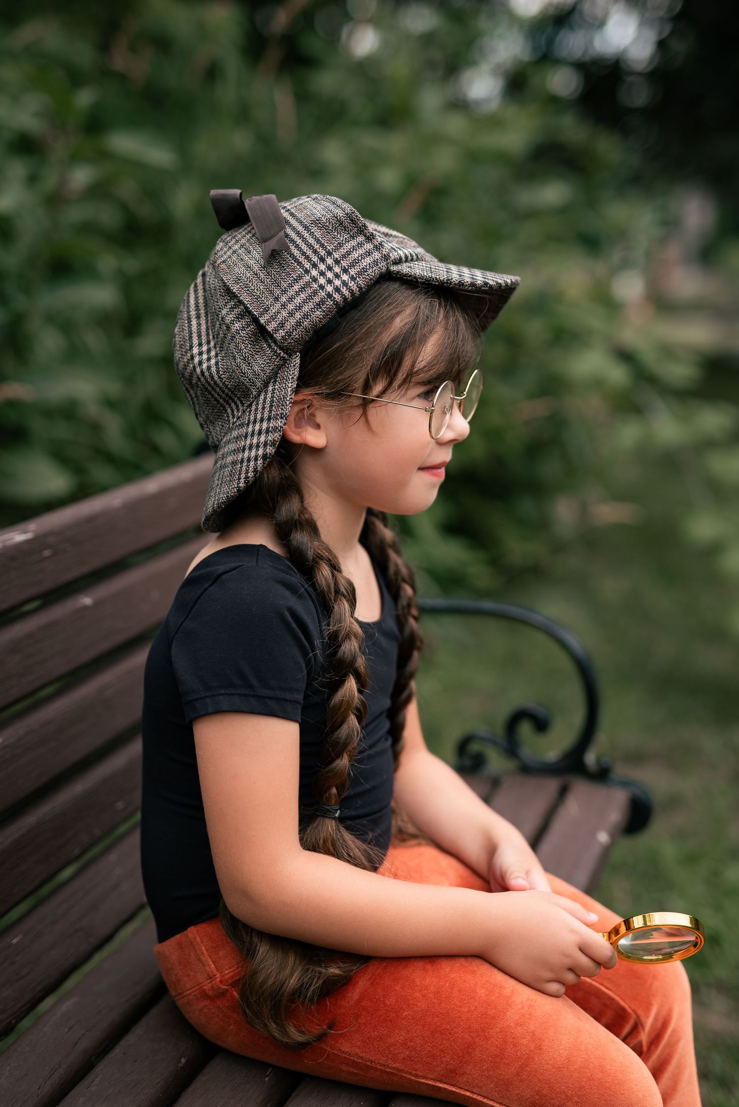 A girl with braids wearing a plaid detective hat and glasses sits on a bench holding a gold magnifying glass outdoors.