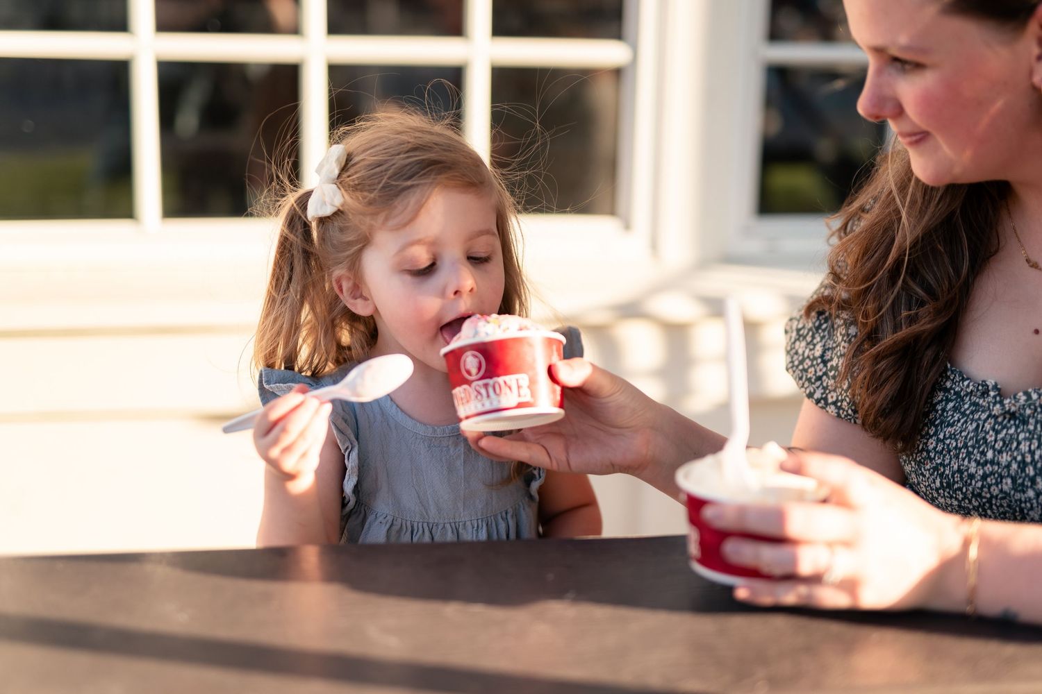 A young girl eagerly eats ice cream from a red cup while a woman holds it for her outdoors.