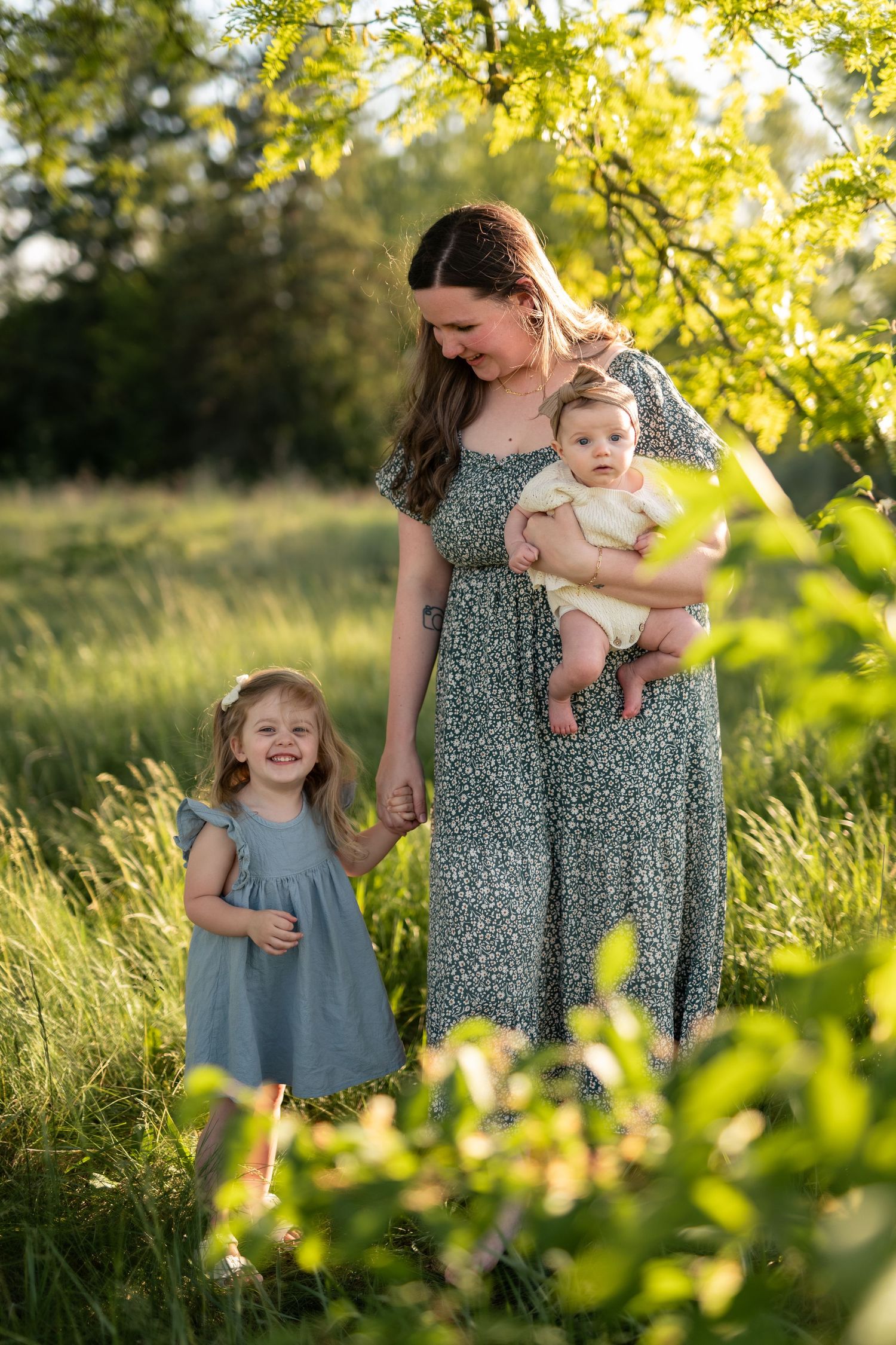 Mother holding baby while toddler daughter holds her hand in sunny green meadow, all wearing blue dresses.