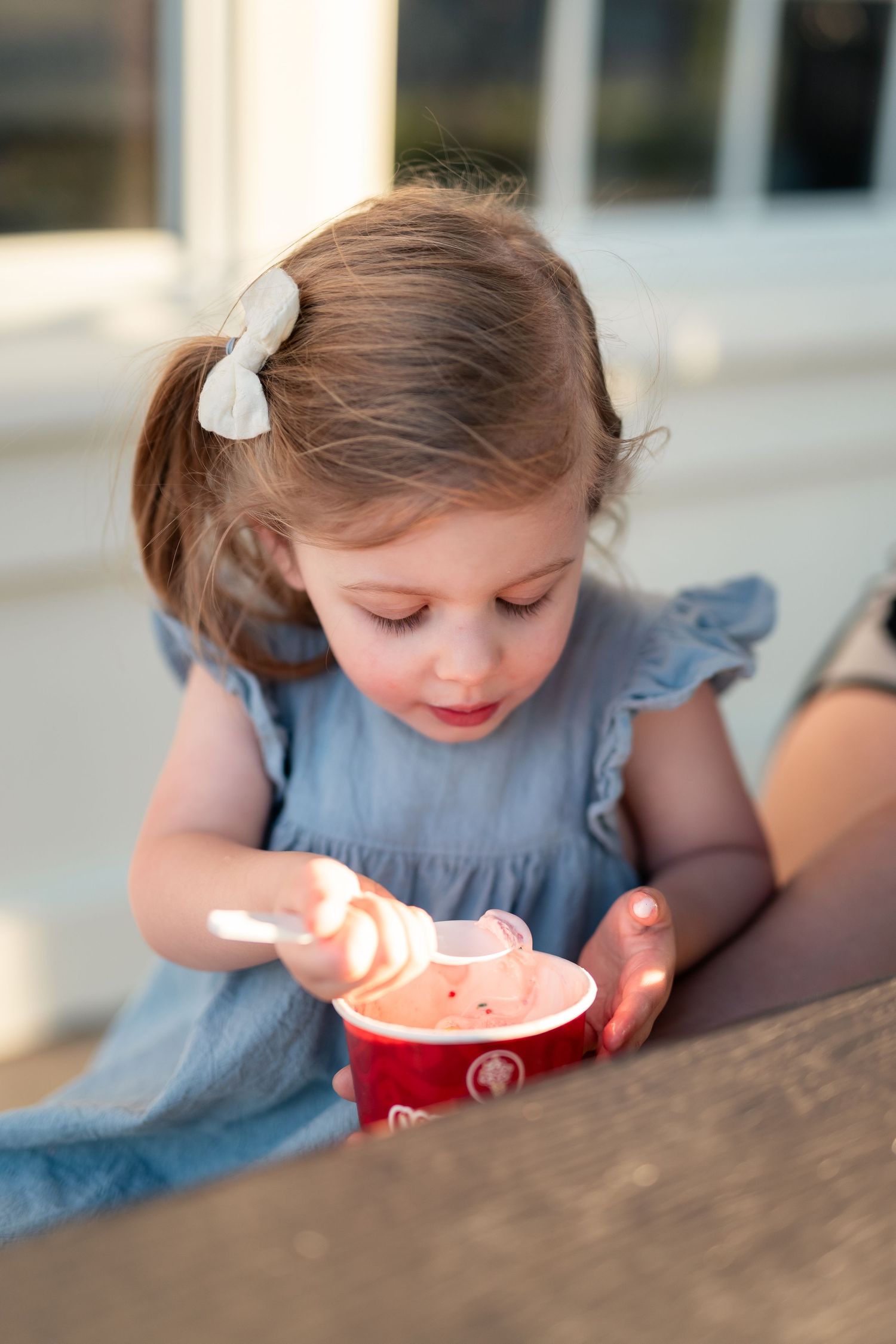 Toddler girl with pigtails eating pink ice cream from a red cup with a spoon outdoors.