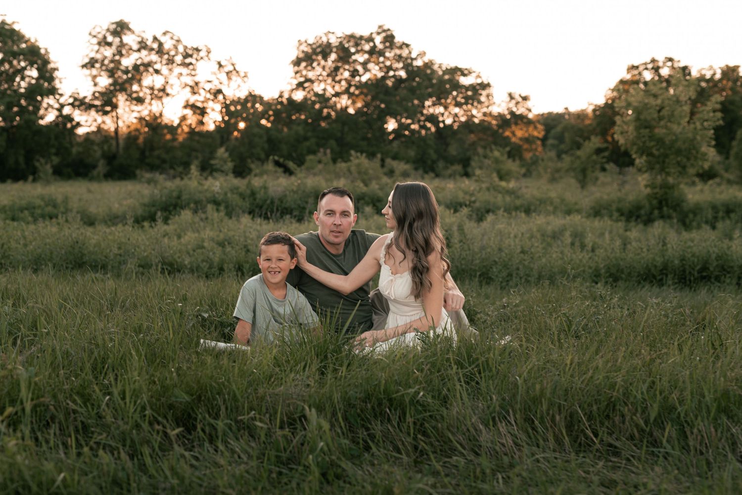 Family of three sitting in a lush green field at golden hour sunset with trees in background.