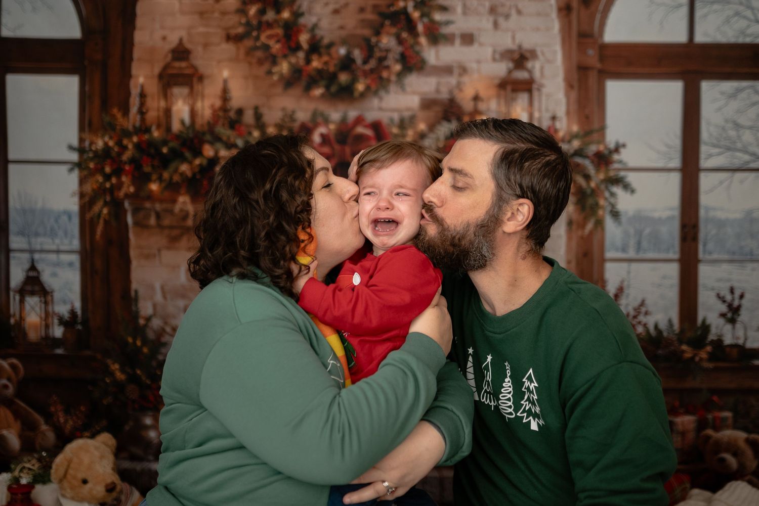 Parents in green sweaters kiss a crying baby in red during a cozy Christmas family photo session with festive wreaths and garland.