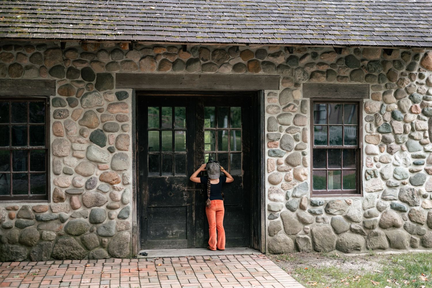 Person in orange pants peering through a black door of a rustic stone building with large rounded rocks.