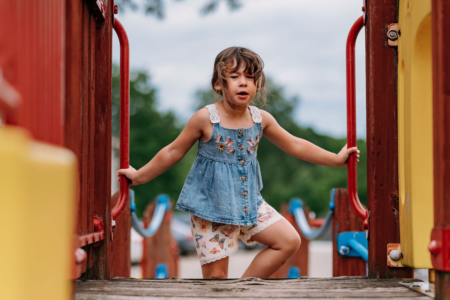 Young child in denim dress playing energetically on red and yellow playground equipment.