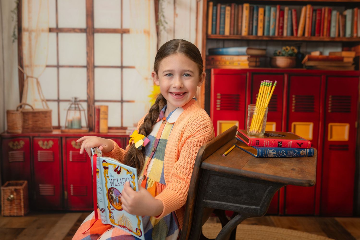 Young girl smiling at desk with book, pencils, and red bookshelf in school-themed photo backdrop.