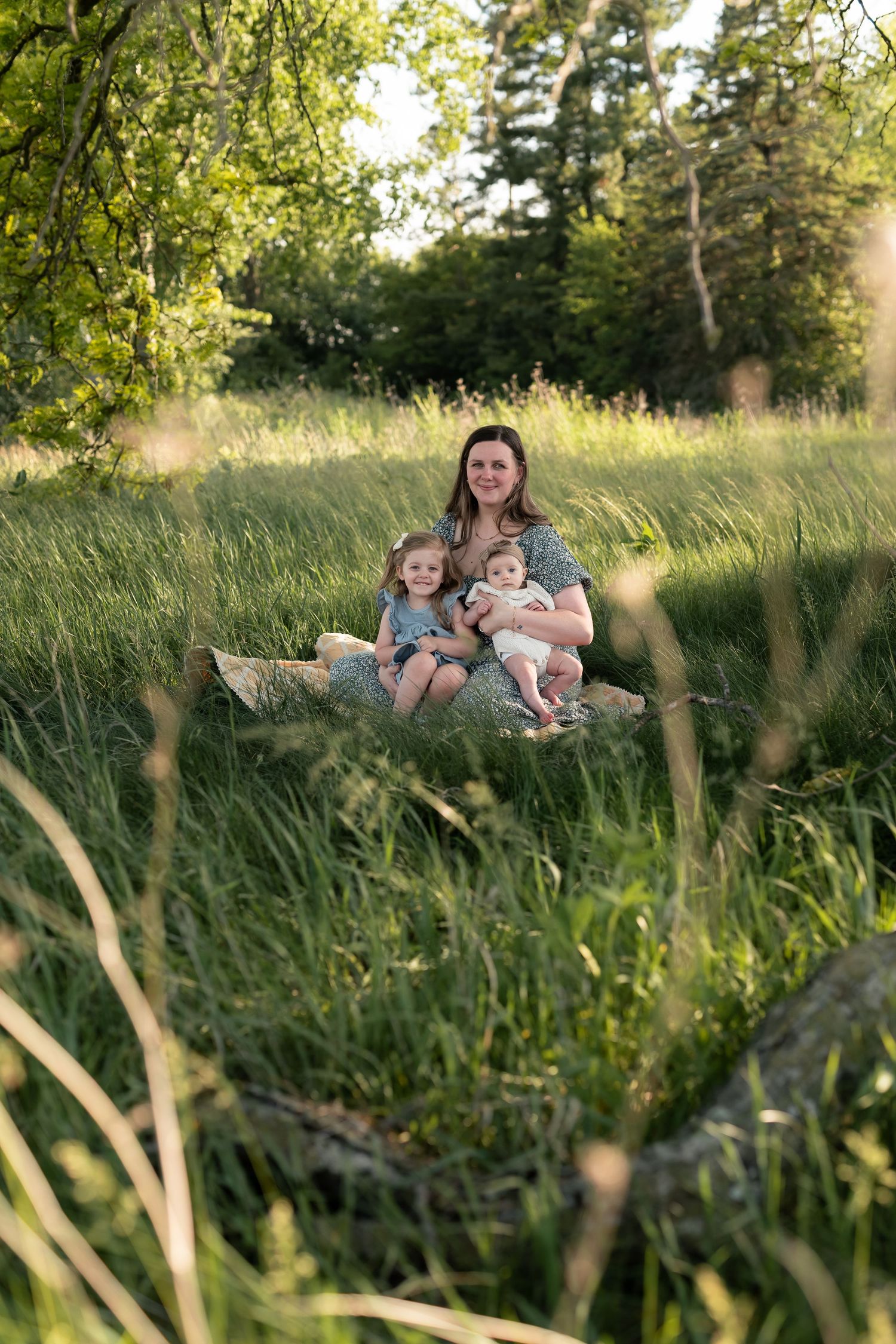 A mother sits with her two young children on a log in a lush green meadow during golden hour.